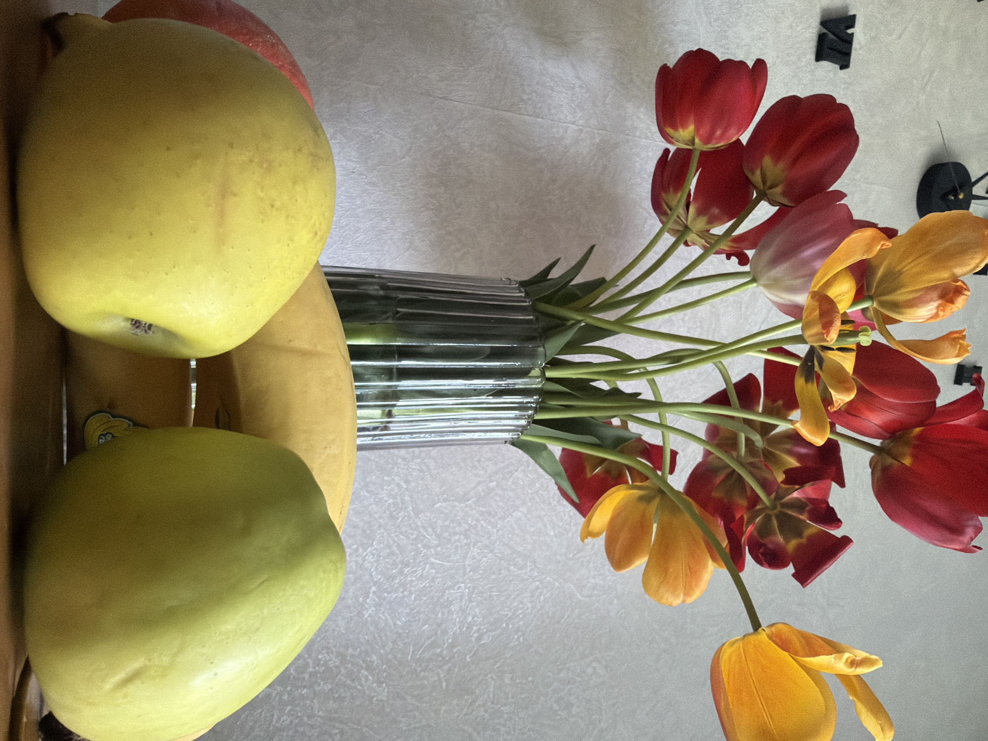 The image shows a still life arrangement, including fruit and a vase of tulips. On the left side are several fruits: two green apples, one yellow apple, and a red apple. The vase is transparent with a ribbed texture and appears to contain water with several green stems of tulips emerging from it. The tulips are a mix of colors, including yellow, red, and orange. A clock is visible in the upper right corner. The background appears to be a textured, neutral-colored surface, possibly a tablecloth. The location in Novosergievka, Russia, is not supported by the image content.