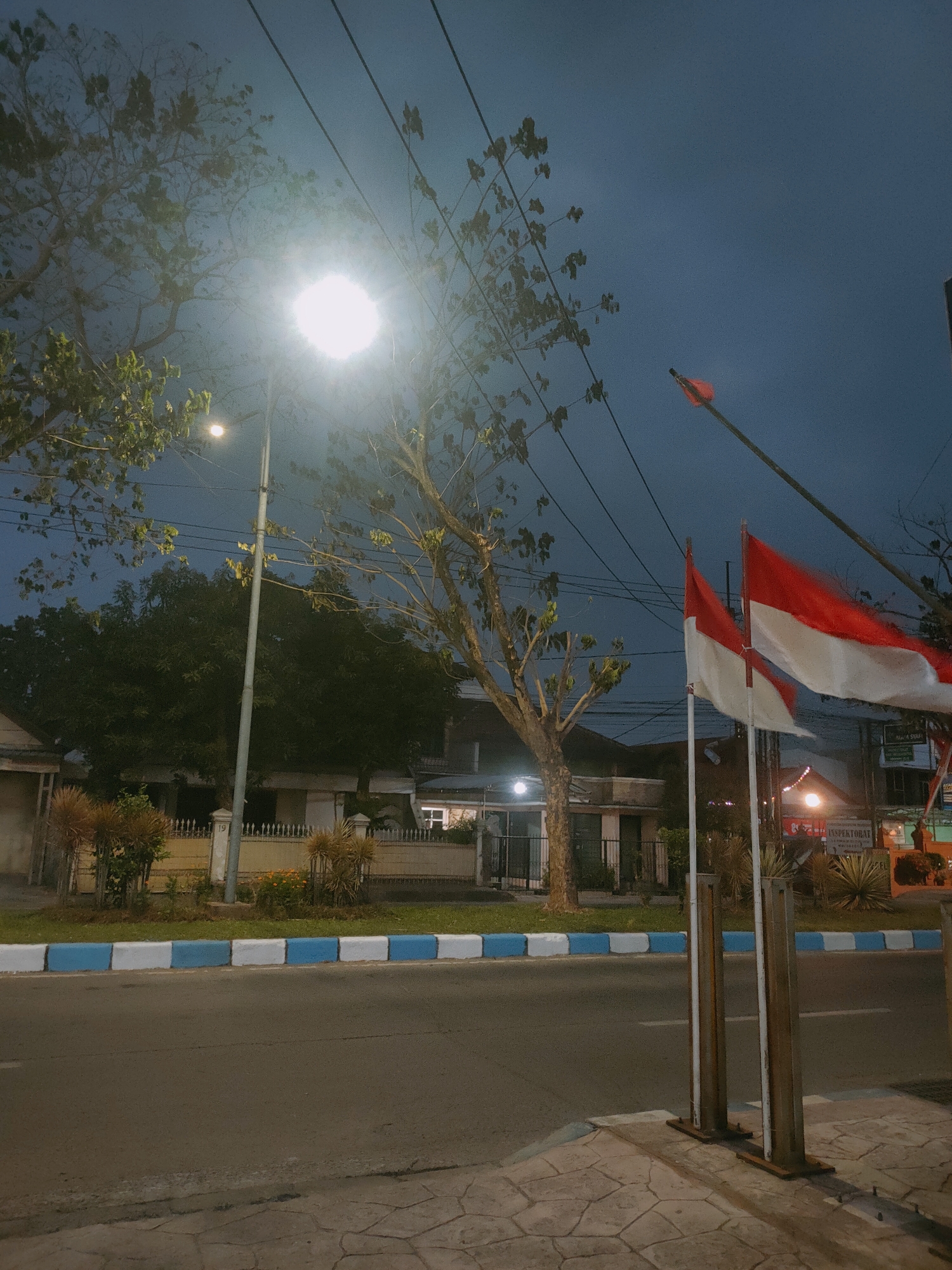 This image depicts a street scene at dusk in Sooko, Indonesia, with two Indonesian flags prominently displayed in the foreground. The flags, red and white, are attached to poles and appear to be waving in the breeze. In the background, a street lamp illuminates the scene, casting a bright glow through the branches of a nearby tree. The street itself has a light gray asphalt surface, bordered by a curb with alternating blue and white painted sections. Beyond the curb, there is a strip of green grass and some bushes, followed by a low-pitched fence and buildings. The sky is a deep blue, characteristic of twilight. Power lines are visible crisscrossing the sky above the trees. The overall impression is that of a quiet street in a residential or commercial area as night begins to fall.
