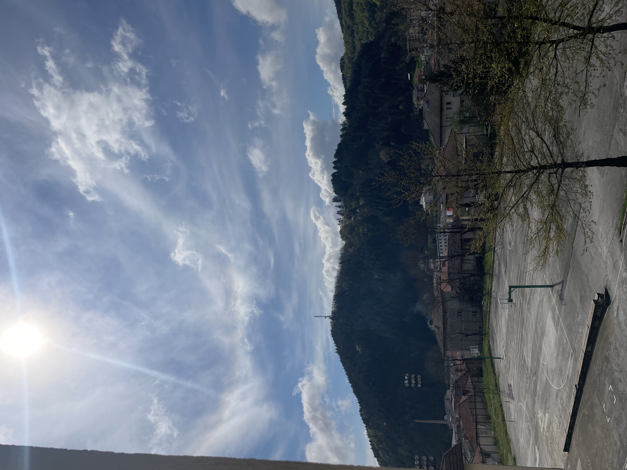 The image displays a panoramic view of a mountainous town under a bright, partially cloudy sky. The scene is illuminated by a prominent sun on the left, which creates a strong lens flare. The sky is a clear blue, adorned with a mix of white, wispy cirrus clouds and some scattered cumulus clouds.
In the middle and background, a large, densely forested mountain range dominates, appearing dark green. A communication tower can be seen atop one of the distant peaks. In the foreground and midground, an outdoor concrete basketball court with clearly marked white lines and two green hoops with backboards is visible. The court is empty. Several bare or newly budding deciduous trees stand near the court, suggesting either early spring or late autumn. Beyond the court and trees, nestled against the base of the mountain, are various buildings, likely residential, characterized by traditional architecture and sloped roofs.
No people or specific activities are depicted, giving the scene a calm and serene atmosphere. The time of day appears to be bright daytime, under clear weather conditions. No visible text is discernible in the image. The setting suggests a town integrated into a natural, hilly landscape.