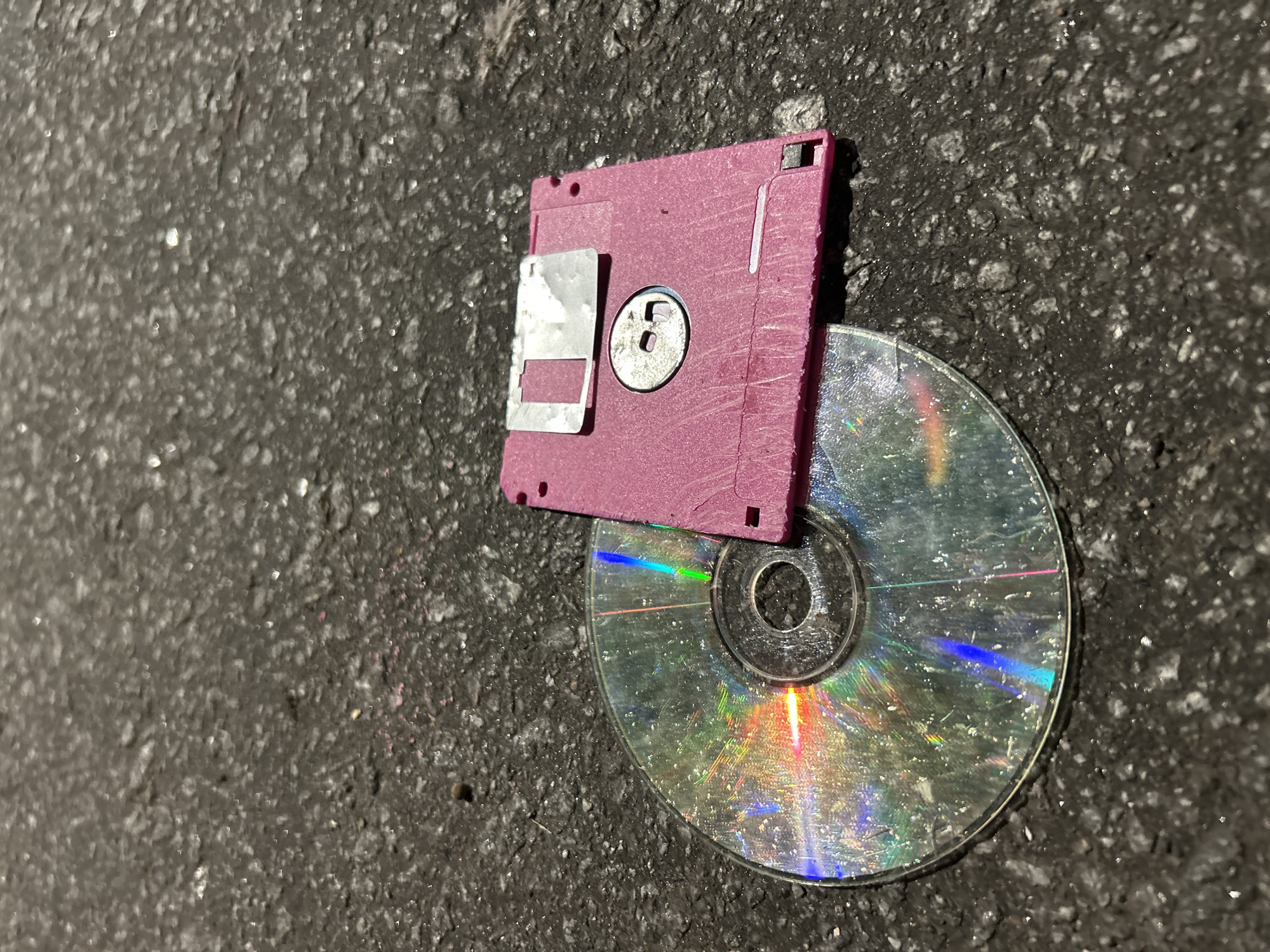 A purple 3.5-inch floppy disk lies atop a scratched and damaged CD on a coarse asphalt surface. The surrounding area is a close-up of the asphalt, with no other discernible features to indicate a specific location in Buenos Aires.