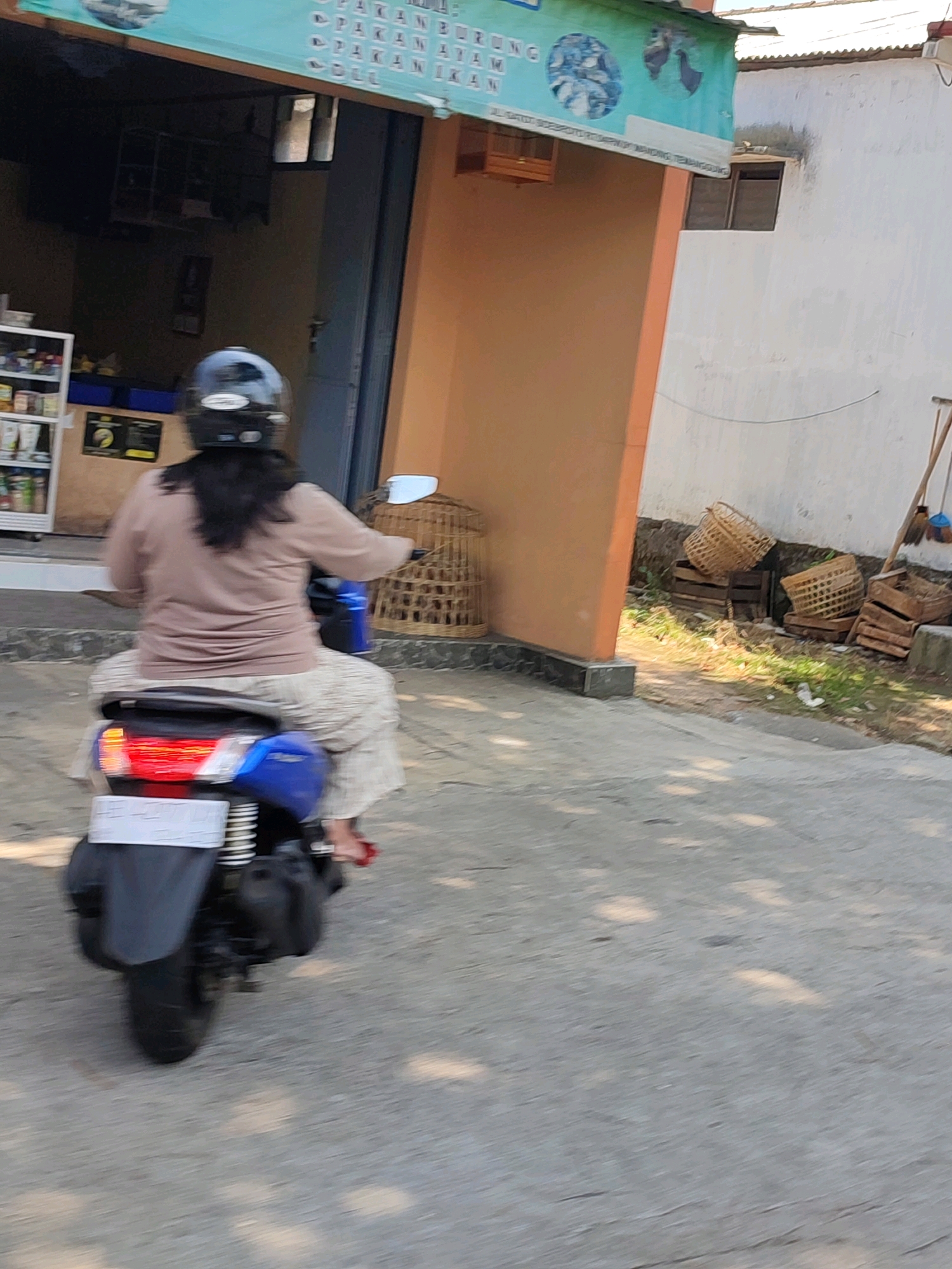 A person wearing a dark helmet and a light brown long-sleeved shirt rides a blue motorcycle. The person is wearing loose-fitting patterned pants and red shoes. The motorcycle has a license plate that is unreadable due to motion blur. The motorcycle is moving on a paved road. In the background, there is a building with an orange facade and a white wall. There are several woven baskets and wooden crates stacked near the white wall. A banner with text and images of birds is visible above the orange facade of the building. This scene is located in Manding, Indonesia, a region known for its craft villages, including the production of woven goods.