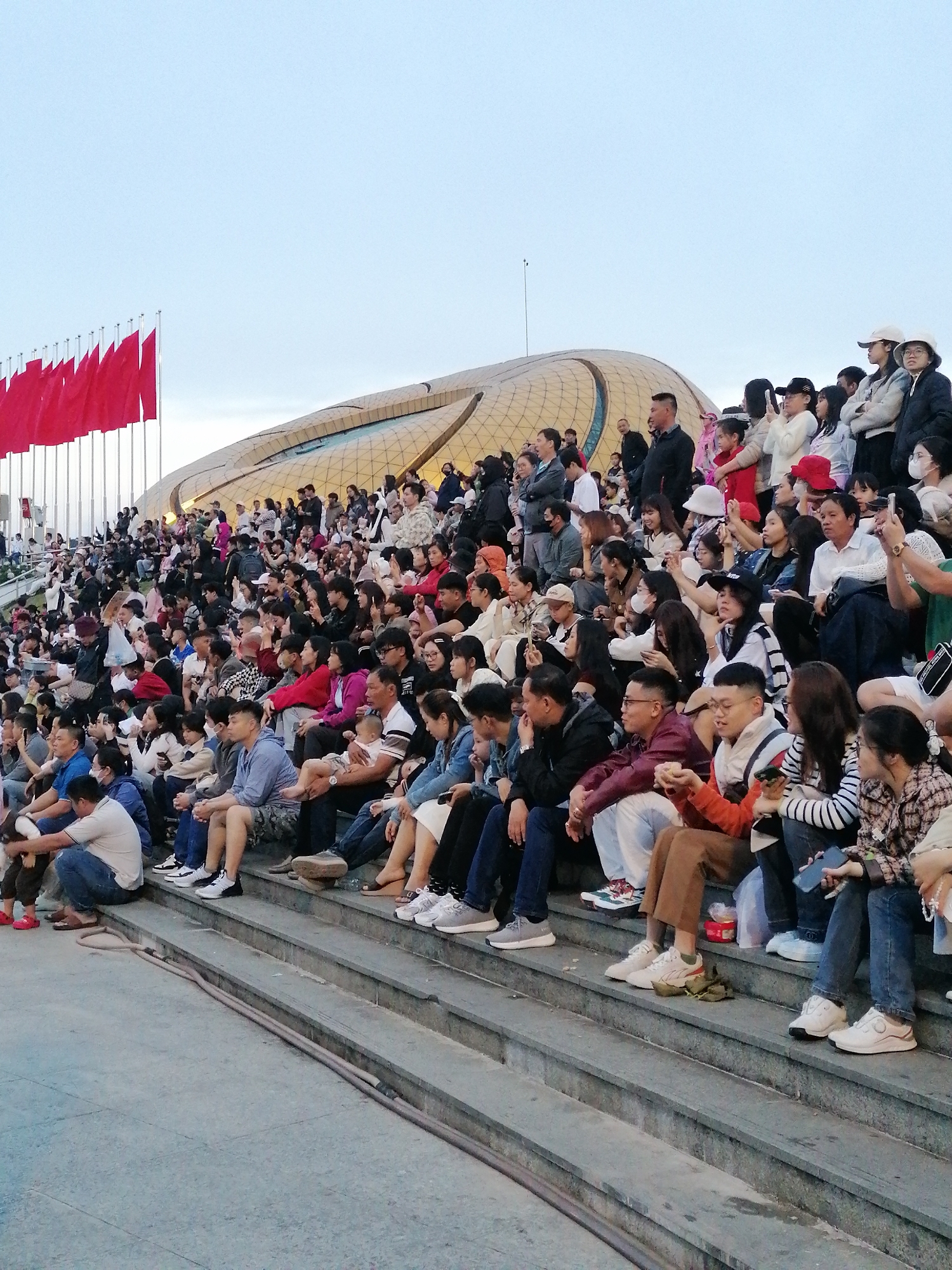 The image captures a large crowd of people seated on outdoor stadium-style steps, facing a stage area that is not fully visible. Red flags are prominently displayed on flagpoles to the left of the frame. In the background, a distinctive, curved, golden building with a patterned facade is visible. The scene appears to be an outdoor event or gathering in Da Lat, Vietnam, likely at a venue such as the Da Lat Palace Convention Center, given the architectural style and the presence of numerous spectators. The people are of various ages, dressed in casual attire, and many are looking in the same direction, suggesting they are watching a performance or presentation. Some individuals are holding up mobile phones, possibly to take photos or videos. The environment is an urban outdoor setting with a clear sky overhead, indicative of daylight.