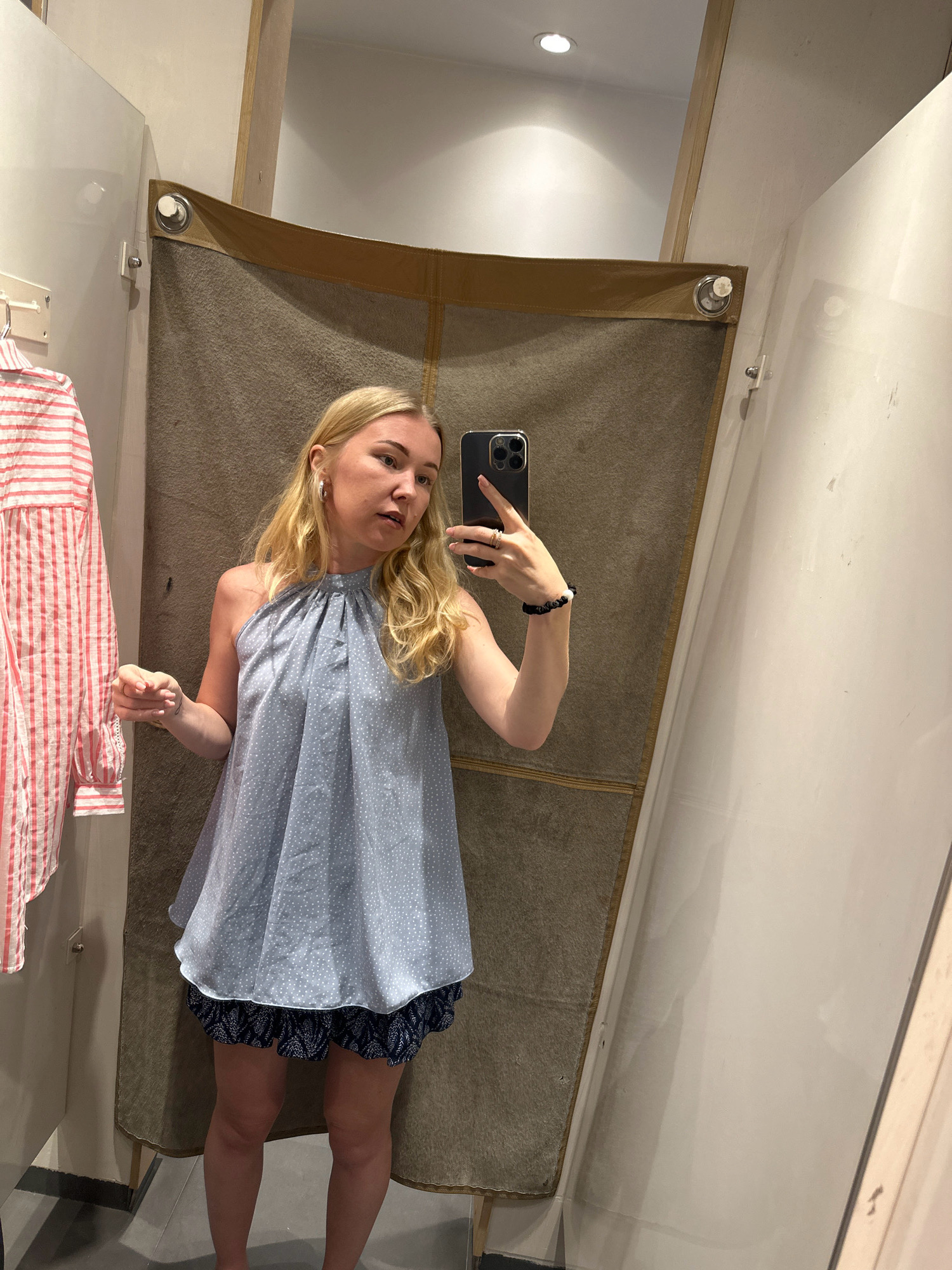 A blonde woman is standing in a fitting room, taking a selfie with her phone. She is wearing a light blue top with white polka dots and dark blue shorts with a pattern. A red and white striped shirt is hanging on a rack to her left. The fitting room has a brown curtain in the background and white walls.