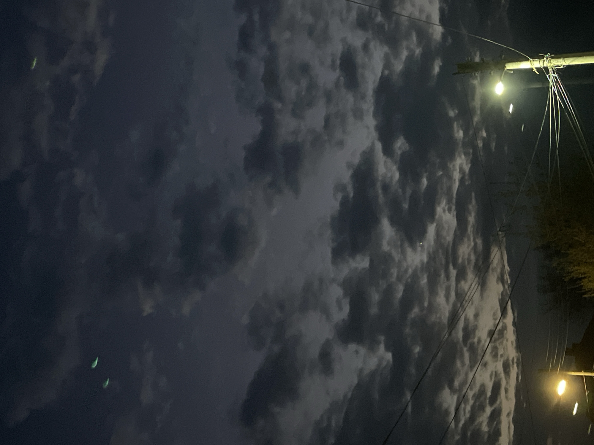 The image shows a night scene in Elhovo, Bulgaria. The sky is filled with dark, heavy clouds. A utility pole with two illuminated streetlights is on the right side. Wires extend from the pole across the sky. A structure or building is partially visible at the lower right edge. Two faintly glowing, green objects are in the lower left part of the sky.
