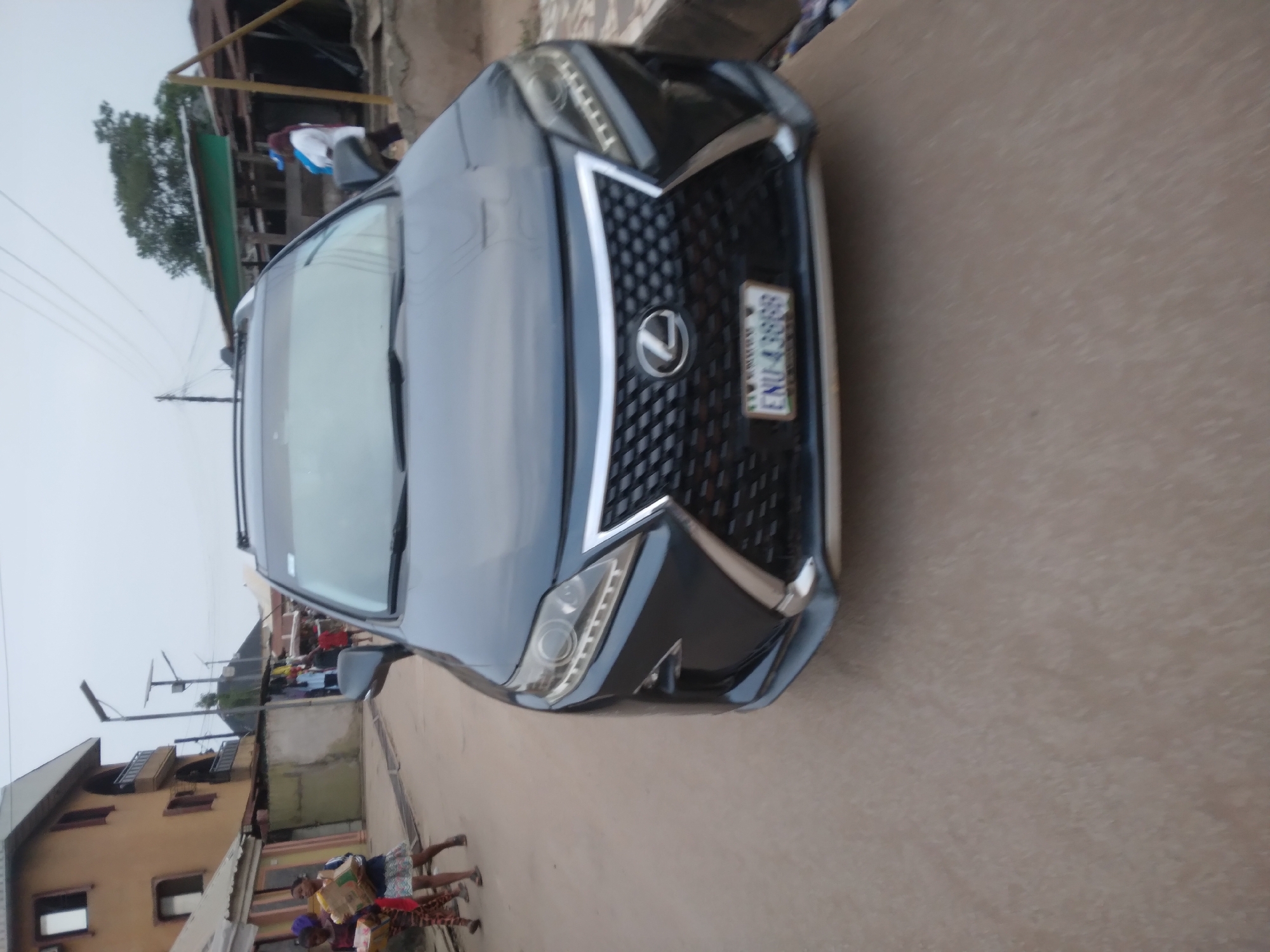The image shows a gray Lexus SUV parked on a paved road in Onitsha, Nigeria. The front of the vehicle is prominently displayed, showing the grille, headlights, and license plate with the inscription "ENU-4388BG". In the background, there are buildings, utility poles, and people walking. The overall setting appears to be a residential or commercial street within the city. The sky is overcast, suggesting it might be early morning or late afternoon.