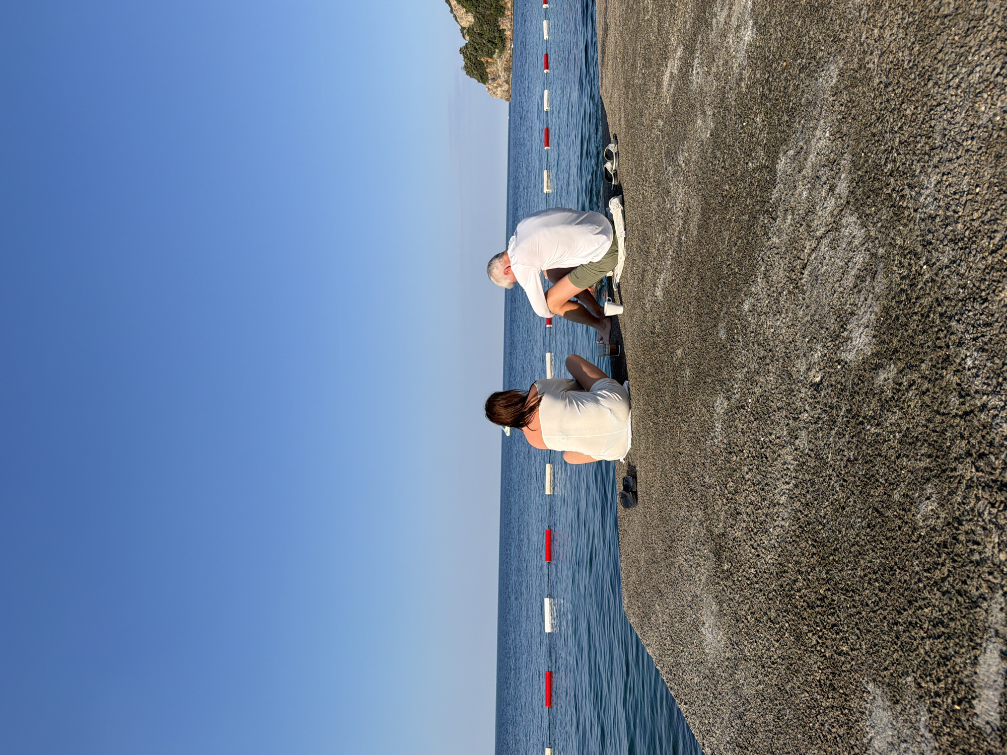 Two individuals, a woman and a man, are seated on a concrete pier overlooking the sea. The woman, with dark hair, wears a white dress and is positioned on the left. The man, with gray hair, is on the right, wearing a white long-sleeved shirt and olive green shorts. Both are facing away from the camera and towards the water. The sea is calm and blue, with a line of red and white buoys in the distance. The sky is clear and bright blue. A rocky, vegetated cliff face is visible on the right side of the frame, in the far distance. On the pier, near the man, are a white mug and a glass, and near the woman are a pair of dark flip-flops. The scene appears to be at a beach or waterfront area in Budva, Montenegro, possibly near Mogren Beach or the Old Town promenade, given the characteristic rocky coastline and clear Adriatic Sea.