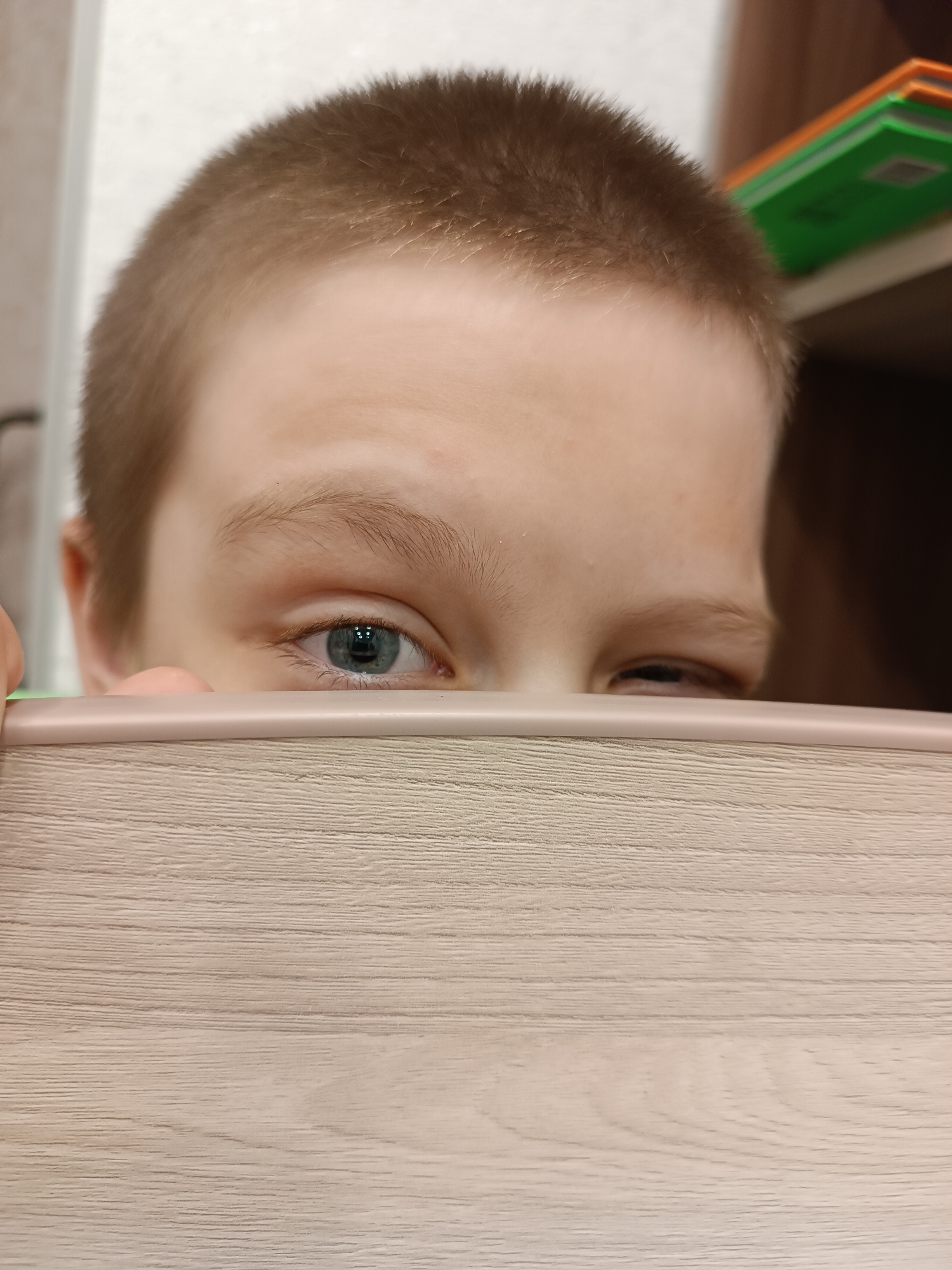 A young boy with short, light-brown hair peeks over the edge of a light-colored wooden surface. His left eye is blue, and he is looking directly at the camera. His right eye appears to be slightly squinted. A portion of his face is visible above the wooden surface, and there are blurred items, potentially books, on a shelf behind him. The image was likely taken indoors. Based on the prompt, this image was likely taken in the city of Chaikovsky, Russia.