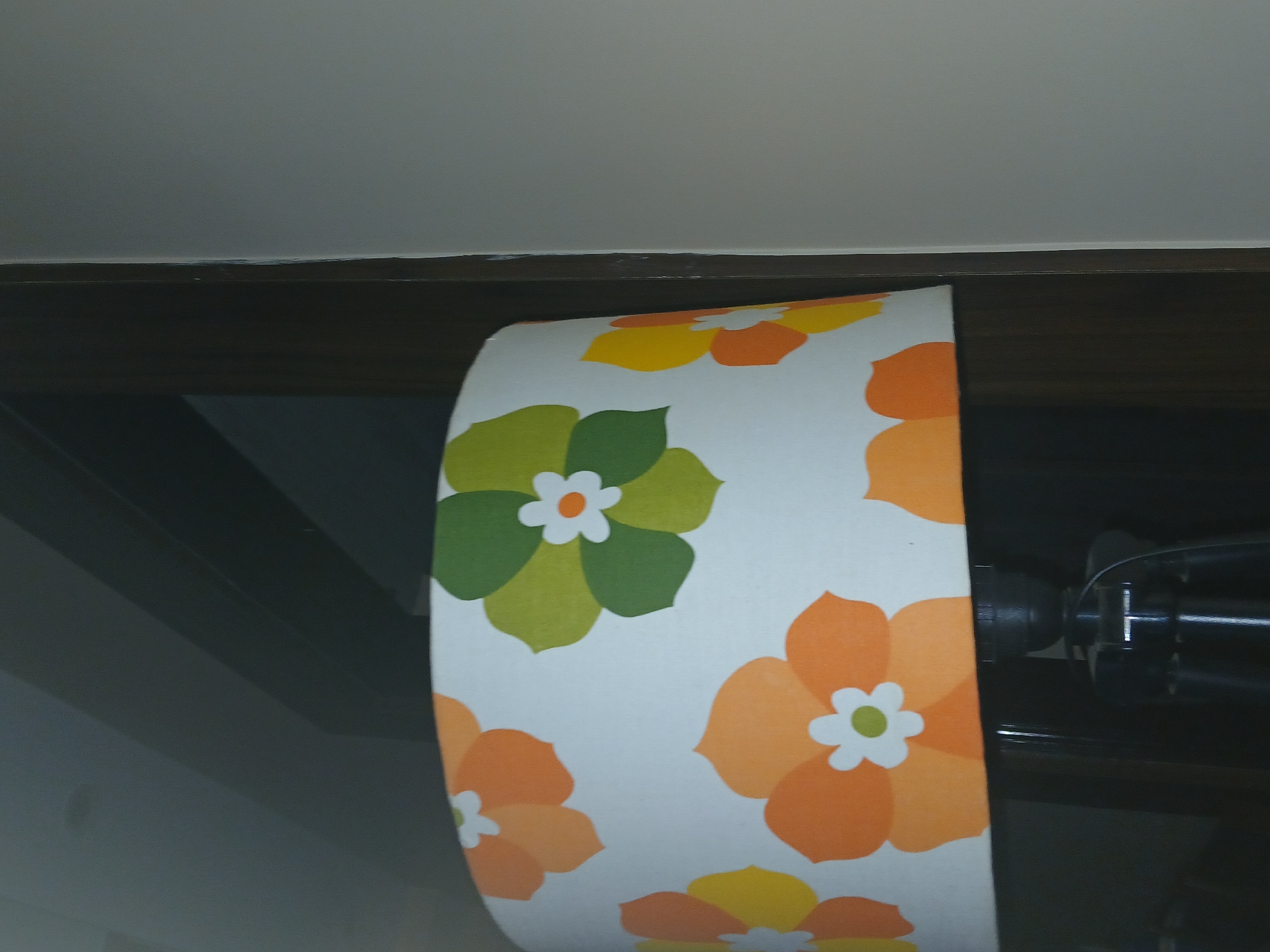 The image shows a section of a floral patterned window roller blind, possibly in a domestic setting in Bengaluru, India. The blind features stylized floral designs in shades of green, orange, and yellow, set against a white background. The blind is partially rolled up, revealing the roller mechanism. The image is taken from a low angle, capturing the ceiling and the upper frame of the window or doorway to which the blind is attached. The overall lighting is subdued.