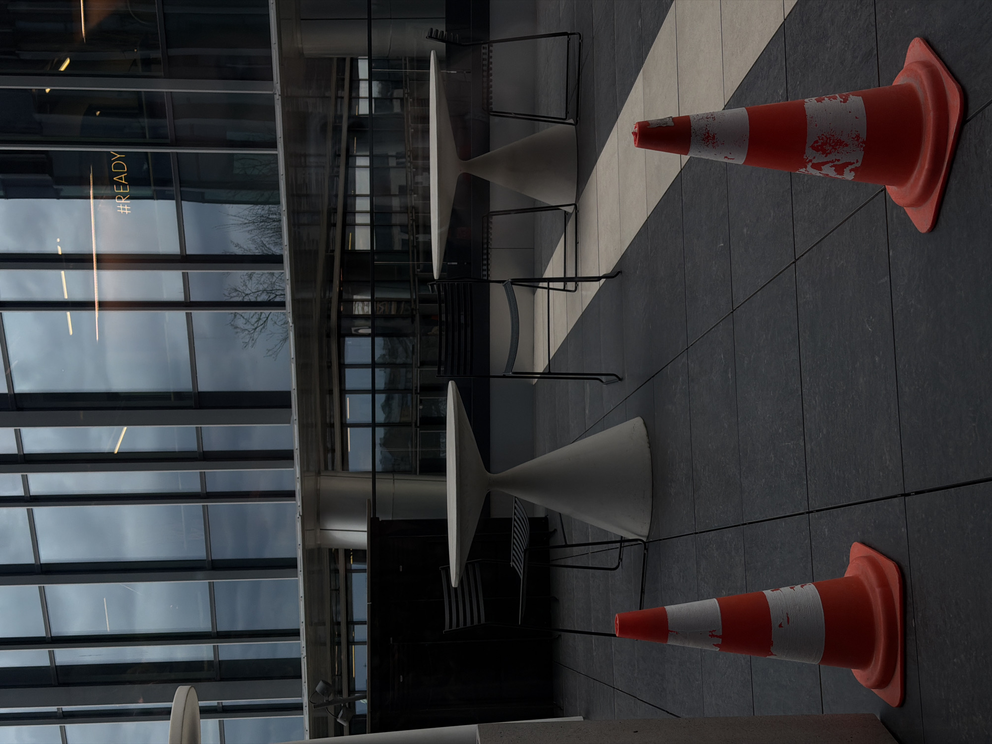 The image depicts an outdoor scene in what appears to be a public space. Architectural elements suggest an urban location. Two orange and white striped traffic cones are present, one in the foreground and one in the background. The cones show signs of wear. Several modern white tables with black metal chairs surround them. The ground is paved with large tiles in contrasting dark and light shades. The windows of a building appear in the background, reflecting the sky. The word "#READY" is visible on a window. Without further evidence it is not possible to specify the precise location in Riga, Latvia.