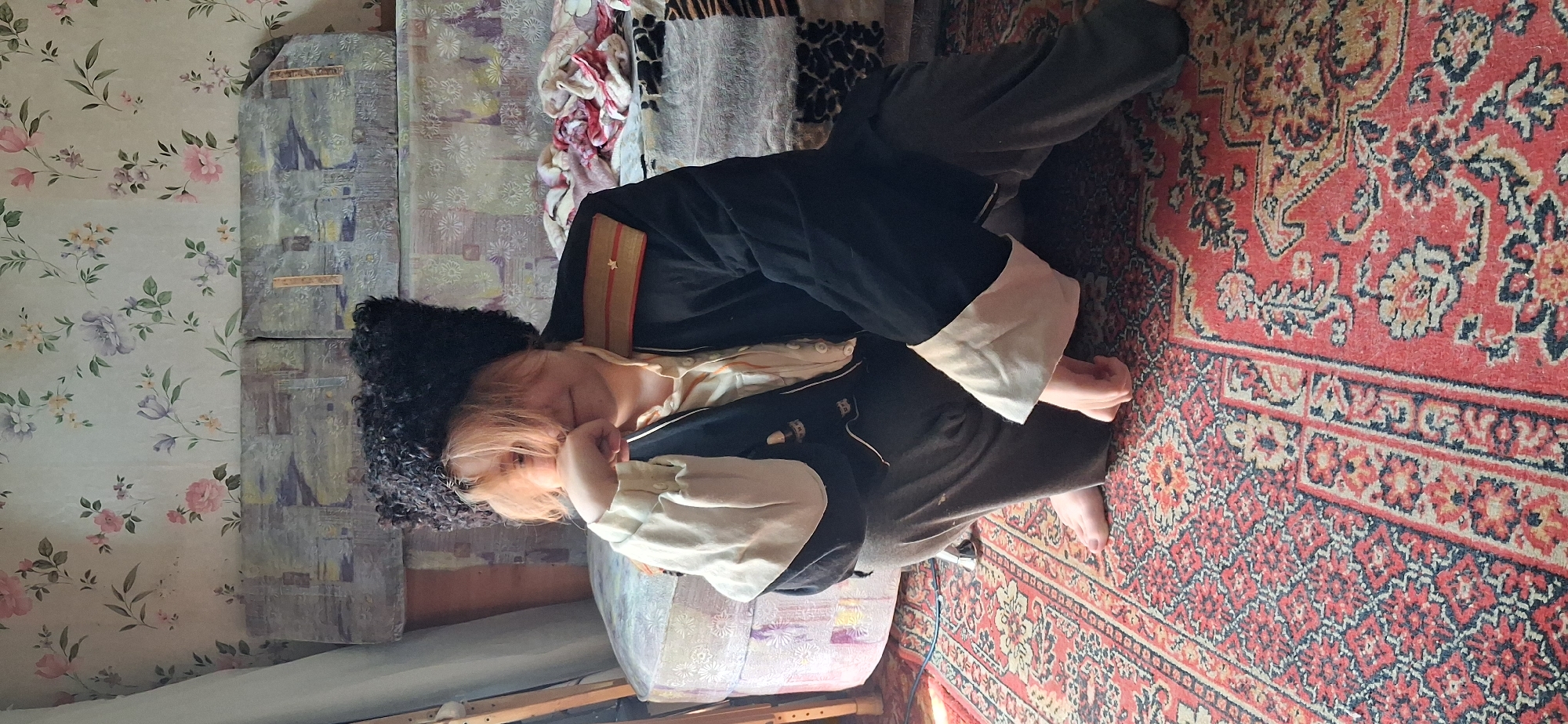 A person wearing a dark, fur-lined hat and a dark jacket with gold trim and a star insignia is kneeling on a patterned rug in an indoor setting. The person has light-colored hair and is resting their chin on their hand, with their gaze directed downwards. They are wearing a light-colored shirt with ruffled cuffs peeking out from the jacket sleeves, and dark pants. Behind the person, a patterned sofa with a floral wallpaper backdrop is visible. The scene appears to be a room with natural light filtering in from the side.
