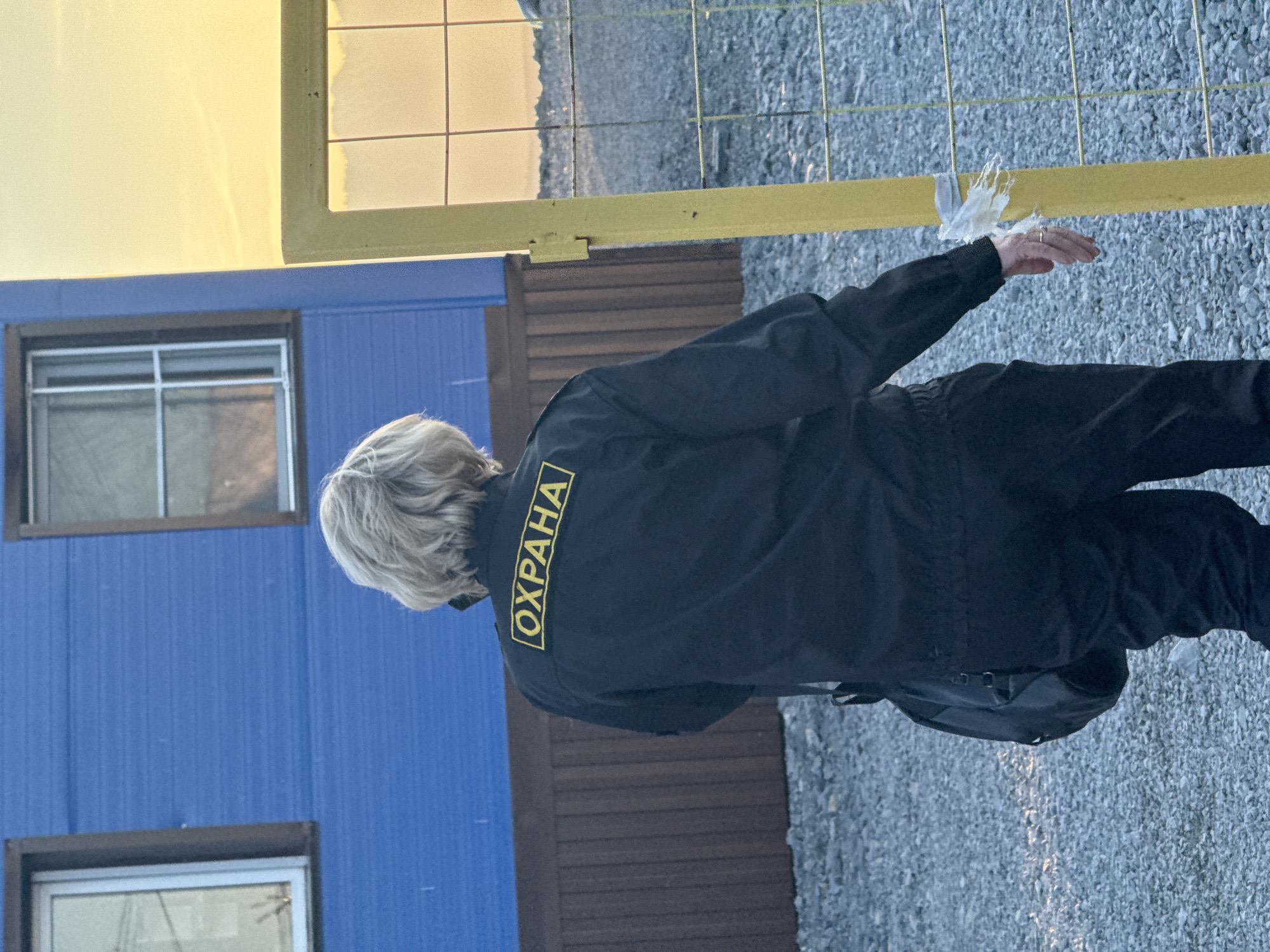 A person wearing a black jacket with the word "ОХРАНА" (meaning "Security" in Russian) on the back stands facing away from the camera. The person has short, light-colored hair. They are wearing dark pants and carrying a dark shoulder bag. In the background, there is a blue building with two windows and a brown wooden fence. To the right, a yellow gate with a wire mesh is partially visible. The ground is covered in small grey stones. The sky in the upper right corner is a pale yellow, suggesting it might be sunrise or sunset. The scene appears to be outdoors in an industrial or utilitarian area.