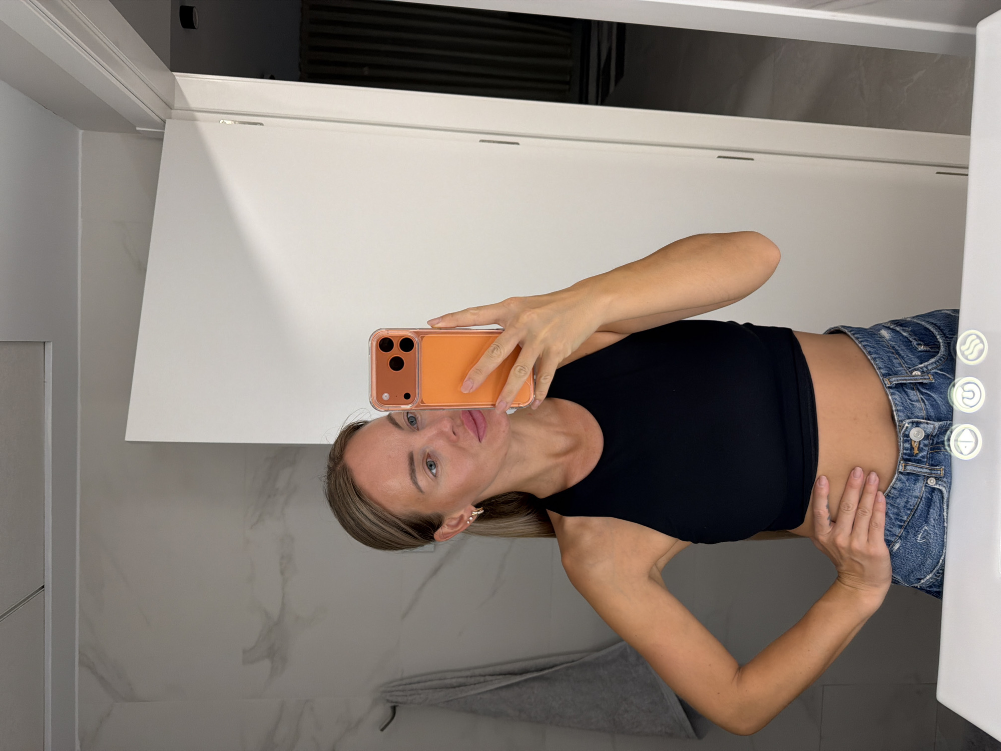 A woman with blonde hair pulled back in a ponytail is taking a mirror selfie in a modern bathroom, possibly located in Finestrat, Spain. She is wearing a black crop top and denim shorts. She is holding an orange-cased smartphone in her right hand, aiming it towards the mirror. Her left hand is placed on her hip. The bathroom features marble-patterned walls, a white vanity, and a closed white door. A grey towel hangs on a rack to the left.