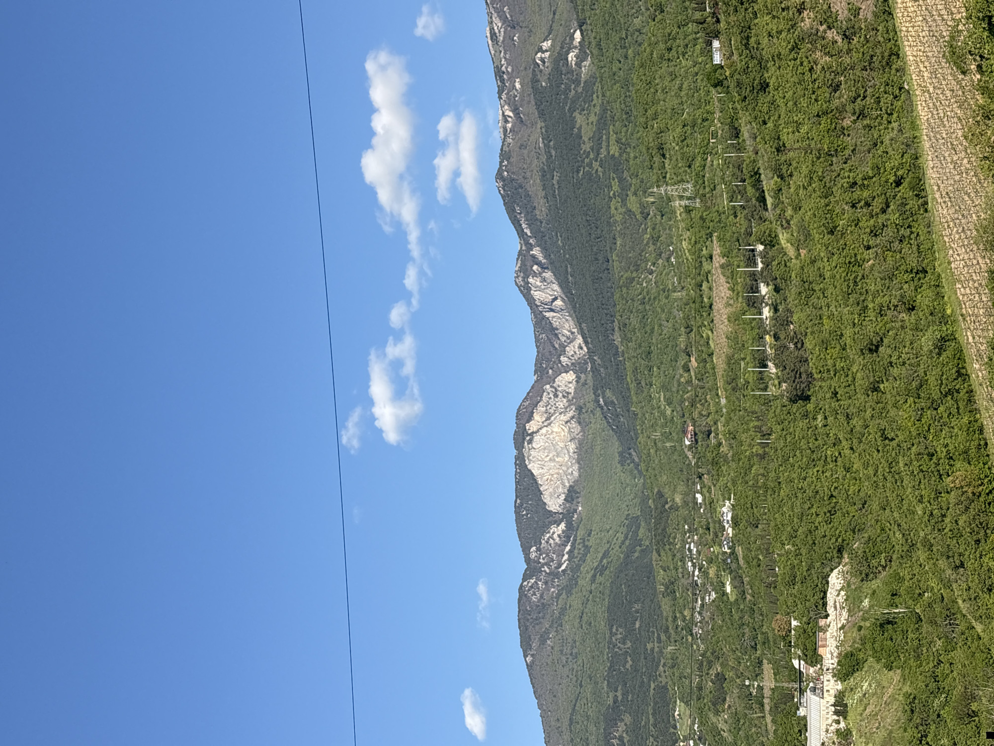 The image shows a mountain range under a bright blue sky with scattered white clouds. The mountains are covered in lush green vegetation, with some rocky, grey cliffs visible. A power line runs across the left side of the image. The location is not in the city of Pushkino, Ukraine. There are no people in the image. The environment is a natural landscape with mountainous terrain and clear skies.