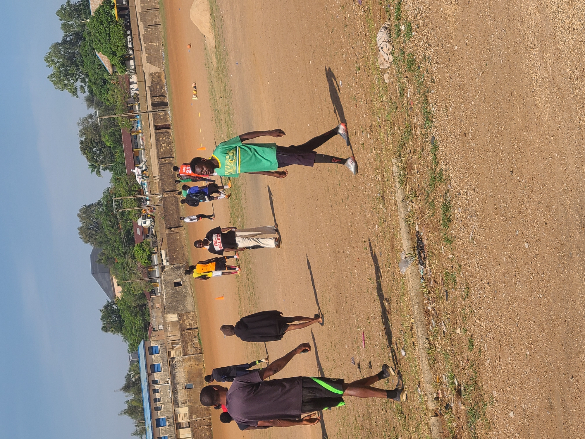 This media file depicts an outdoor sports training session on a reddish-brown dirt field in Jos, Nigeria. The clear blue sky and long shadows cast by the individuals indicate it is either early morning or late afternoon.
The primary subjects are a group of approximately ten to twelve men and boys engaged in what appears to be a football or general fitness practice. They are dressed in athletic attire, including t-shirts, shorts, and tracksuits. Some individuals are standing, while others appear to be walking or jogging across the field. Training aids such as orange cones are visible on the ground, and a small orange ball can be seen in the distance.
Notable details include:
* A person in the mid-ground wearing a green t-shirt and black shorts with "Coop." partially visible on the leg.
* Another individual wearing a black t-shirt with white lettering that reads "REA COLLECT".
* A person in a red jersey with the number "23" is partially visible in the left mid-ground.
* A person in a dark blue or black t-shirt and shorts with green side stripes is in the foreground, facing away from the camera.
The setting includes a strip of grass and some scattered debris along the right edge of the field. In the background, there are various buildings, some with blue accents, and a dense line of green trees. A white truck or utility vehicle is also visible among the buildings. The scene conveys an active community or school sports environment.