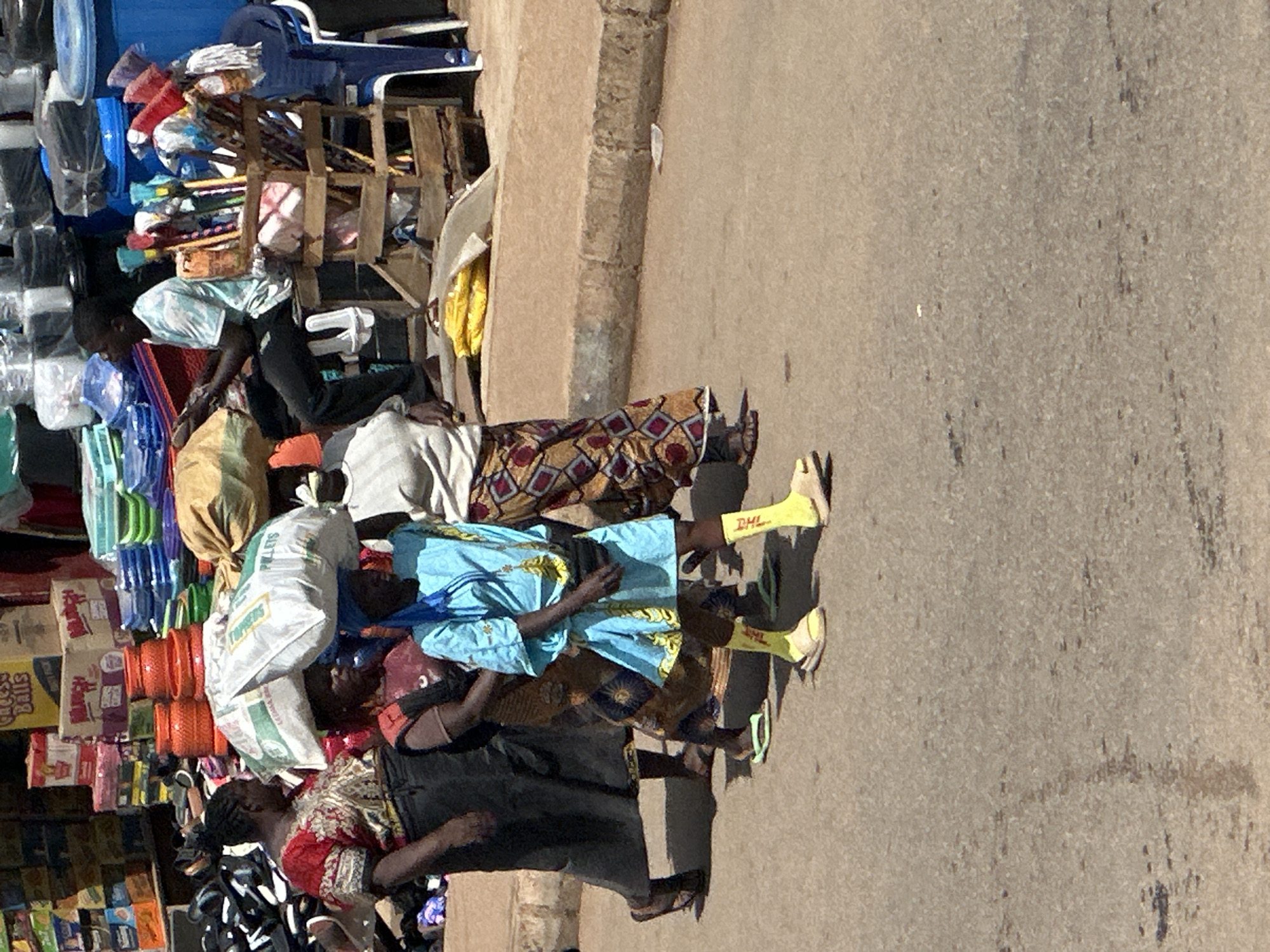 In the city of Bukuru, Nigeria, a street scene unfolds with a group of individuals walking on an asphalt road. In the background, various goods are displayed for sale, including stacked boxes of food items like "Cheese Balls" and "Perritos," as well as colorful plastic containers. To the left, a woman in a red and patterned outfit carries a large white bag on her head. Beside her, two other women, one in a dark top and patterned skirt, and another in a blue dress with yellow trim and bright yellow socks, also walk along. In the middle ground, a young man sits on a wooden stool, appearing to be engaged in a transaction involving a collection of brooms, mops, and cleaning supplies displayed in a wooden crate. The environment suggests a bustling market or roadside stall area.