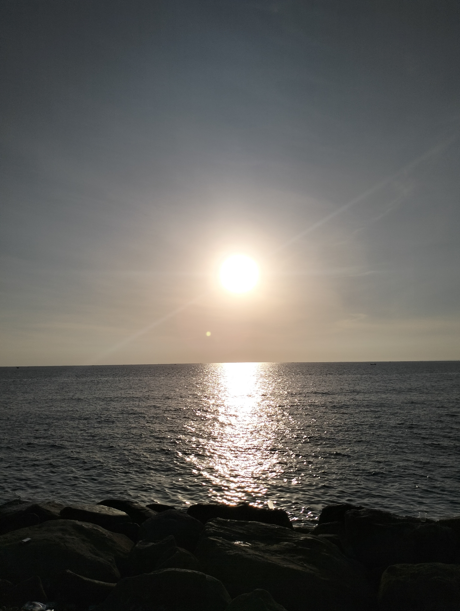 The image shows a seascape at sunset. The sun is positioned high in the sky, reflecting intensely on the ocean surface. The foreground consists of dark rocks. The sky is a gradient of light gray, and the water is a dark gray color with bright reflections of the sun. A few small boats are visible on the horizon. The location is likely a coastal area in the city of Lhokseumawe, Indonesia.