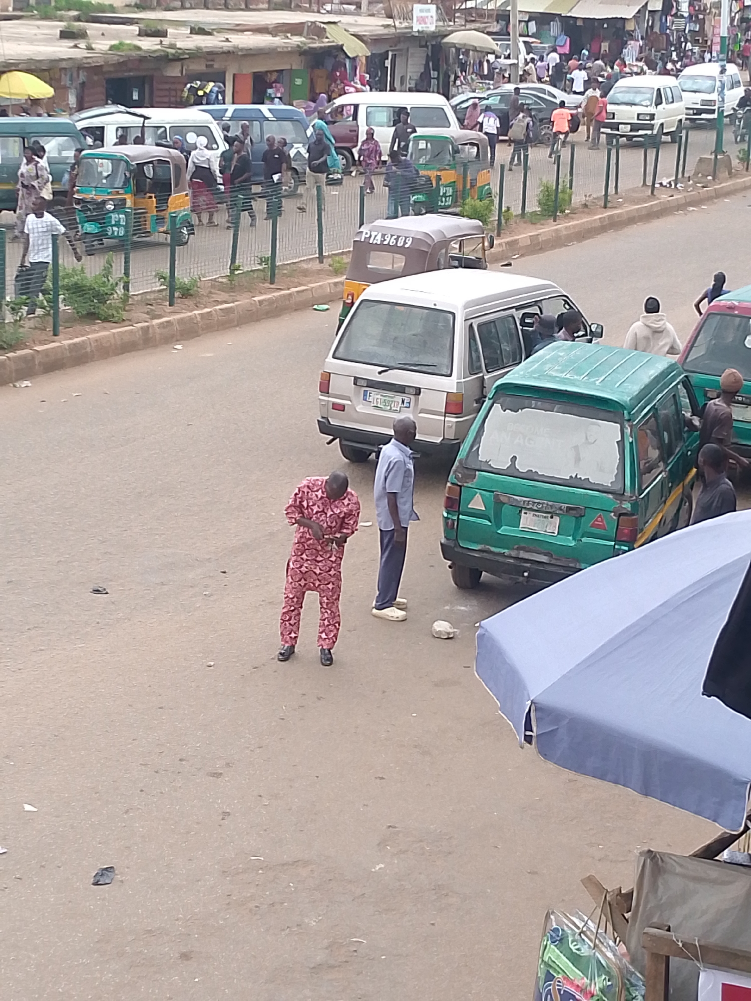 The image depicts a busy street scene in Bukuru, Nigeria, likely a commercial area. In the foreground, a man wearing a pink and red patterned traditional attire is standing in the middle of the road, looking down at his hands. To his right, another man in a light blue shirt and dark pants stands facing the side. A green van with a sticker on the rear window that reads "BECOME AN AGENT" is parked on the right side of the road. Other vehicles, including a silver van and various motorcycles and tricycles (keke napep), are visible on the road and sidewalk. The background shows a row of shops and stalls with people milling about, suggesting a marketplace. The overall atmosphere is one of daily commerce and transportation in a Nigerian urban setting.