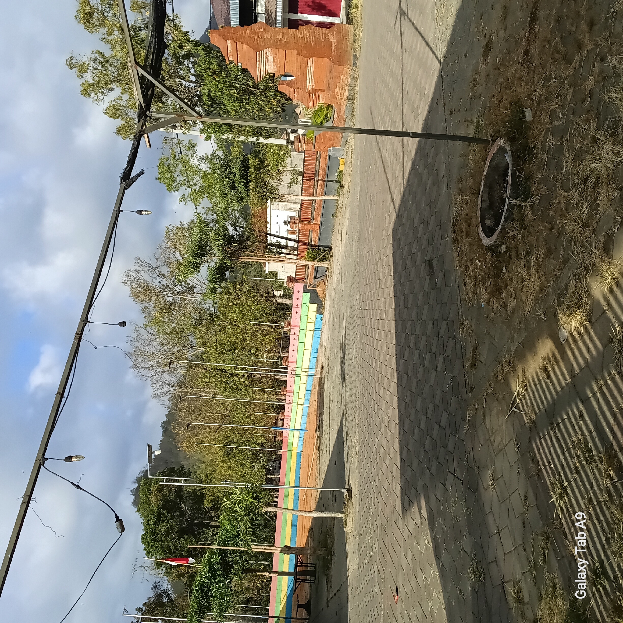 The image shows an outdoor paved area with a fire pit in the foreground. A colorful tiered seating area is visible in the background, along with trees, a brick structure resembling a gate, and buildings. A flag is also visible. The sky is partly cloudy. The location is identified as Bendorejo, Indonesia. There are no people visible in the image.