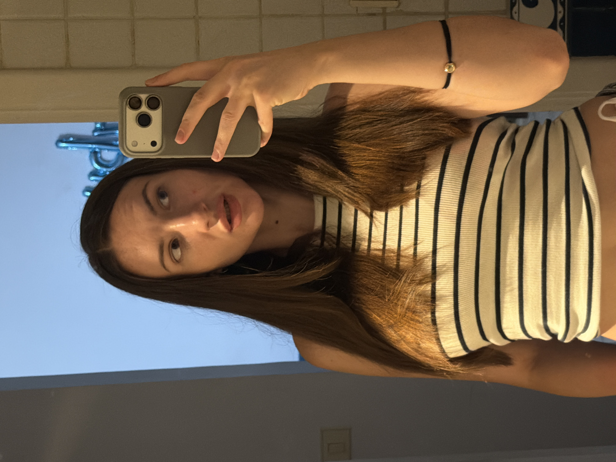 A young woman with long, dark brown hair is taking a selfie in a bathroom. She is wearing a striped tank top and has a black bracelet with a gold bead on her right wrist. She is holding a gray smartphone in her right hand to take the picture. The background shows a tiled wall and a light blue surface with what appears to be metallic blue balloon letters. The environment is likely a residential bathroom. There is no indication of the specific location in Sharm El Sheikh, Egypt.
