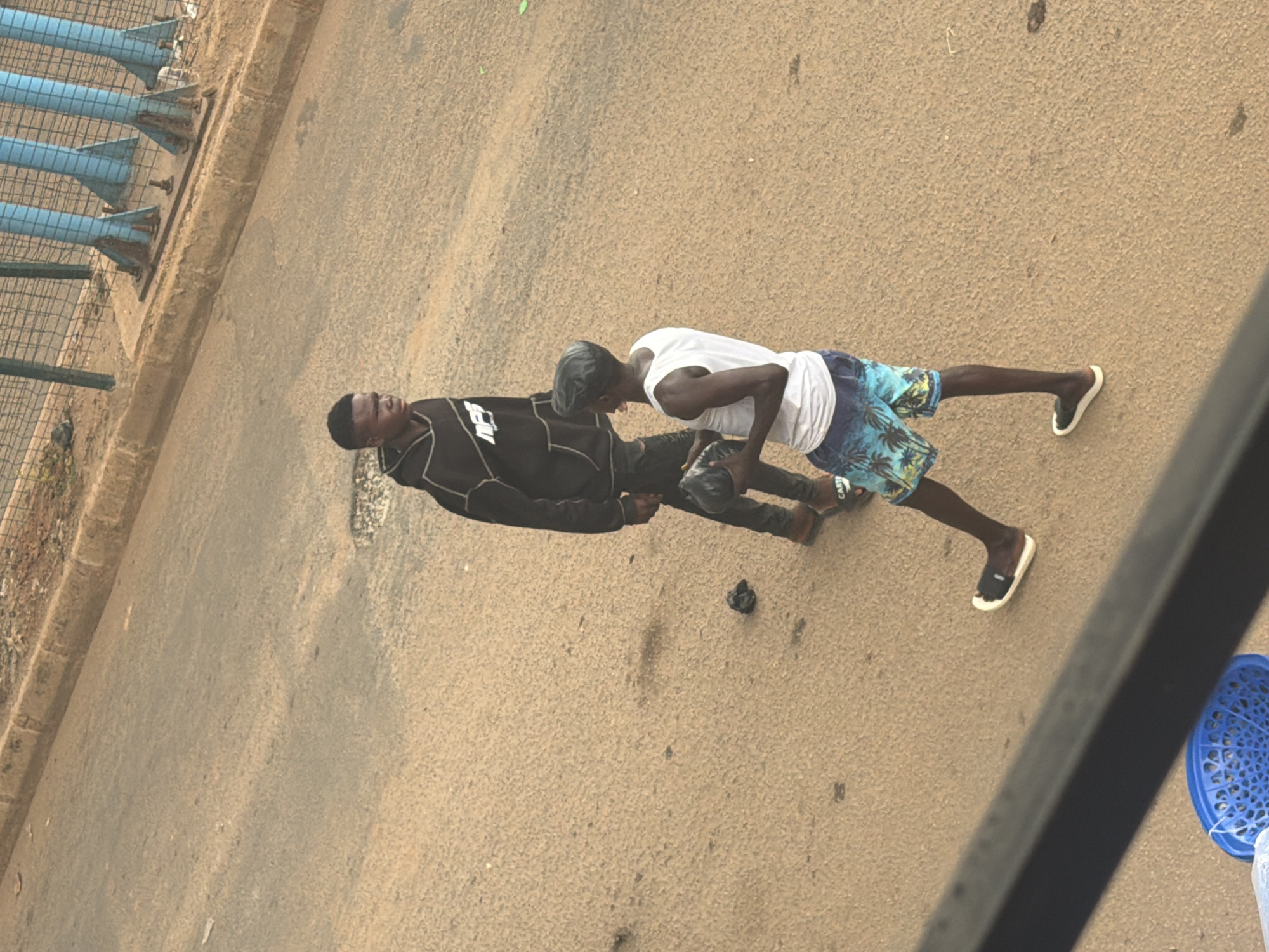 Two young men are walking on a dusty street, likely in Bukuru, Nigeria. The man on the left wears a black hooded sweatshirt with a white graphic on the front and black pants. He is looking to his left. The man on the right wears a white tank top and colorful patterned shorts. He is looking down and appears to be holding a dark object, possibly a hat or a bag, in his hands. Both men are wearing sandals. The street surface is rough and appears to be unpaved or poorly maintained, with some debris scattered on it. In the background, there is a fence and blue vertical structures, suggesting the presence of infrastructure. A blue plastic basket is visible in the foreground, partially obscuring the view of the street.