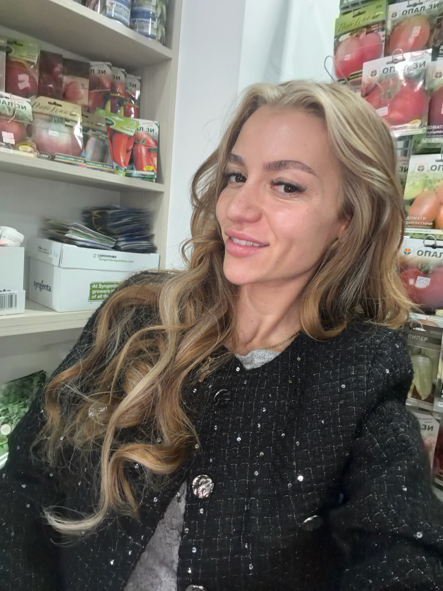 In the city of Sandanski, Bulgaria, a woman with blonde hair is posing for a photo in what appears to be a retail store, possibly a gardening or agricultural supply shop. She is wearing a black sparkly jacket over a gray top and is looking towards the camera with a smile. Behind her, shelves are filled with packets of seeds, predominantly for vegetables like tomatoes and peppers, indicated by the text on the packaging. Some boxes from Syngenta are also visible, suggesting a commercial environment. The lighting is bright, likely from overhead store lights.