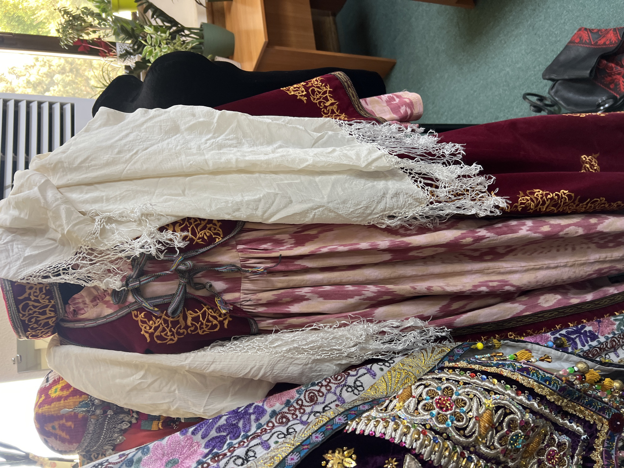 The image shows a close-up of several traditional garments displayed on mannequins or hangers in what appears to be an indoor setting. The foreground features a richly embroidered purple vest with intricate beadwork and gold thread, worn over a pink and white ikat-patterned dress. A cream-colored shawl with white lace trim is draped over the garments. To the left, another garment with floral embroidery in purple and pink is visible. In the background, a red velvet robe with gold embroidery is visible, as well as a dark-colored fabric that could be part of another outfit or a backdrop. The environment includes a window with greenery and sunlight filtering through, a wooden desk, and a section of a green carpeted floor.