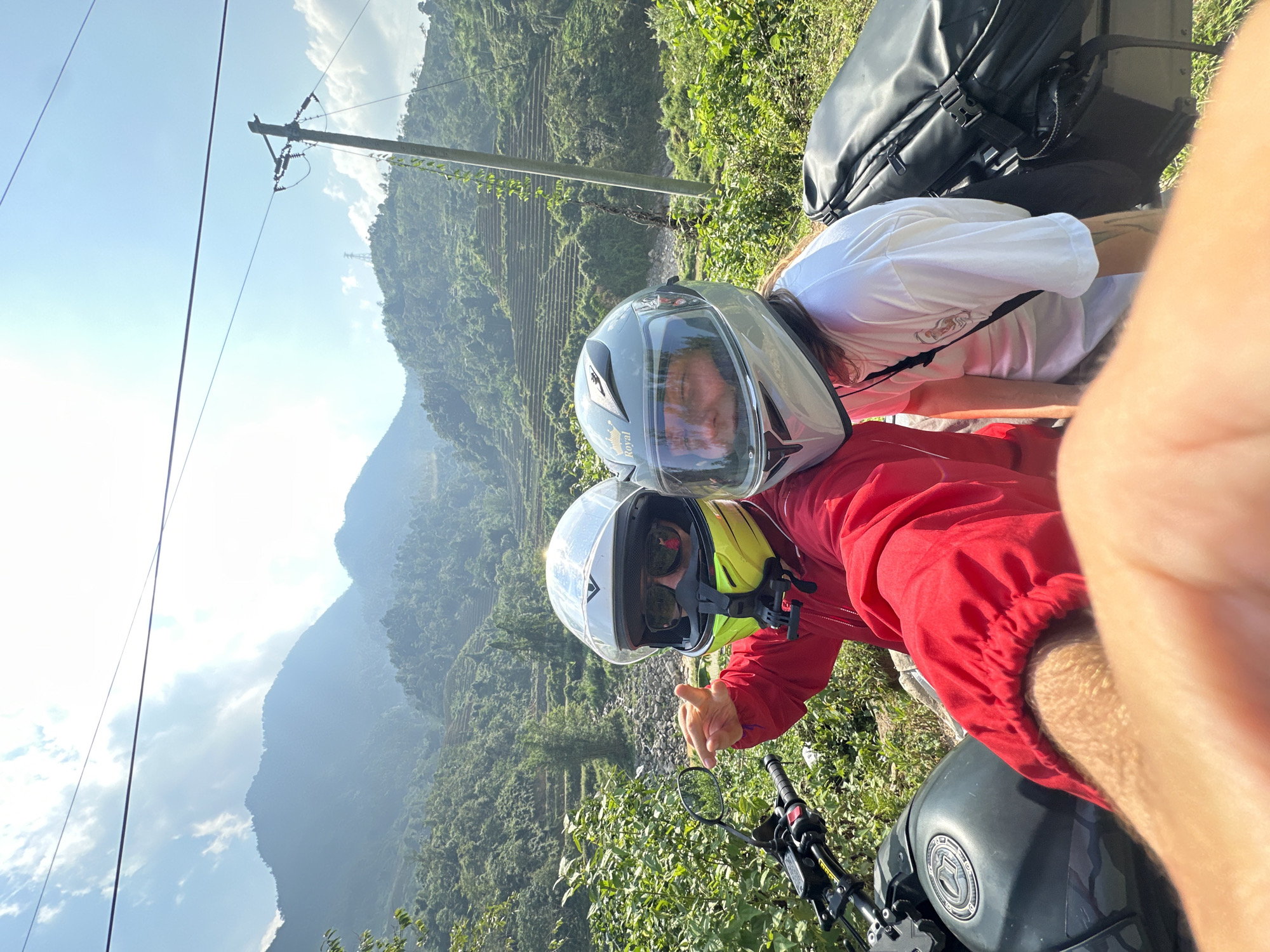 This image captures a selfie of two individuals on a motorcycle, set against a stunning mountainous backdrop, indicative of Thông Nguyên Commune, Vietnam. The photograph appears to have been taken with the camera held by the person in the foreground, resulting in a slightly tilted perspective.
The person in the foreground is wearing a vibrant red jacket and a light-colored motorcycle helmet with a clear visor. Through the visor, sunglasses are visible, and the individual appears to be smiling or making a positive gesture with their right hand, part of which is visible. Directly behind them is another person, dressed in a white t-shirt, also wearing a grey motorcycle helmet with "Royal" written on the side. Their face is visible through the helmet's visor, and long hair can be seen cascading from beneath the helmet.
Parts of a black motorcycle are visible in the lower foreground, including the handlebars, a rearview mirror, and a section of the fuel tank. A large black backpack is strapped to the rear of the motorcycle, suggesting a journey or trip.
The background reveals majestic, verdant mountains covered in dense foliage. Distinct terraced hillsides, characteristic of agricultural practices in the region, are prominent. A wooden utility pole with power lines cuts across the left side of the frame, contrasting with the natural landscape. The sky is bright blue with scattered white clouds, indicating clear, sunny weather, likely during the daytime. Both individuals appear to be enjoying their ride through the scenic, green landscape.