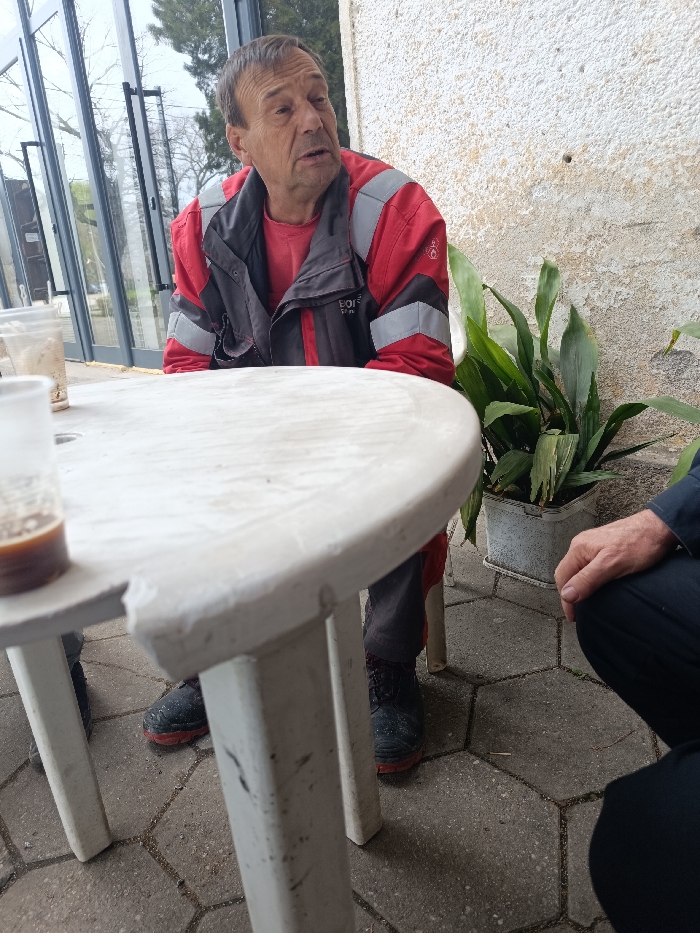 The image presents an outdoor setting in Vinitsa, Bulgaria. An elderly male with short brown hair is seated, oriented right, his mouth slightly open, indicating speech. He wears a red long-sleeved shirt and a red, grey, and black work jacket with reflective banding. Dark work boots with red soles are visible on his feet, resting on hexagonal grey paver tiles. In the foreground, a weathered white plastic round table occupies the central and left portions of the frame. Two plastic cups are on the table, with one in the extreme foreground containing a dark liquid. A second individual's hand and dark clothing are partially visible on the lower right, positioned low to the ground, suggesting a squatting or kneeling posture. The background on the left features large glass panels reflecting trees and an overcast sky. On the right, a textured, light-colored wall with visible wear and a small dark hole serves as a backdrop, with a potted green plant in a rectangular container situated against it.