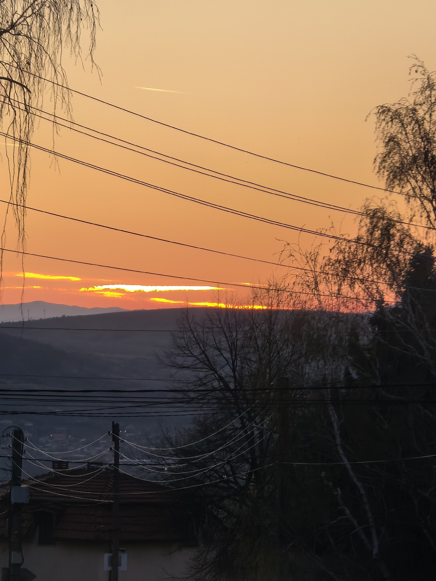 The image captures a vibrant sunset over a hilly landscape in what appears to be a residential area. The sky is a gradient of orange, with the sun partially obscured by a layer of clouds on the horizon, creating a bright yellow glow. The hills are silhouetted against the sky. Power lines crisscross the foreground, and bare trees frame the view on either side. Below the horizon, there is a suggestion of buildings or rooftops in the distance, indicating a town or city. In the very foreground, the roof of a building with a tile texture is visible, along with a utility pole. The location appears to be on the outskirts of a city, as indicated by the mix of natural and built environments. The scene is static, showing no people or animals, and the primary interaction is between the natural elements of the landscape and the artificial structures of the residential area.