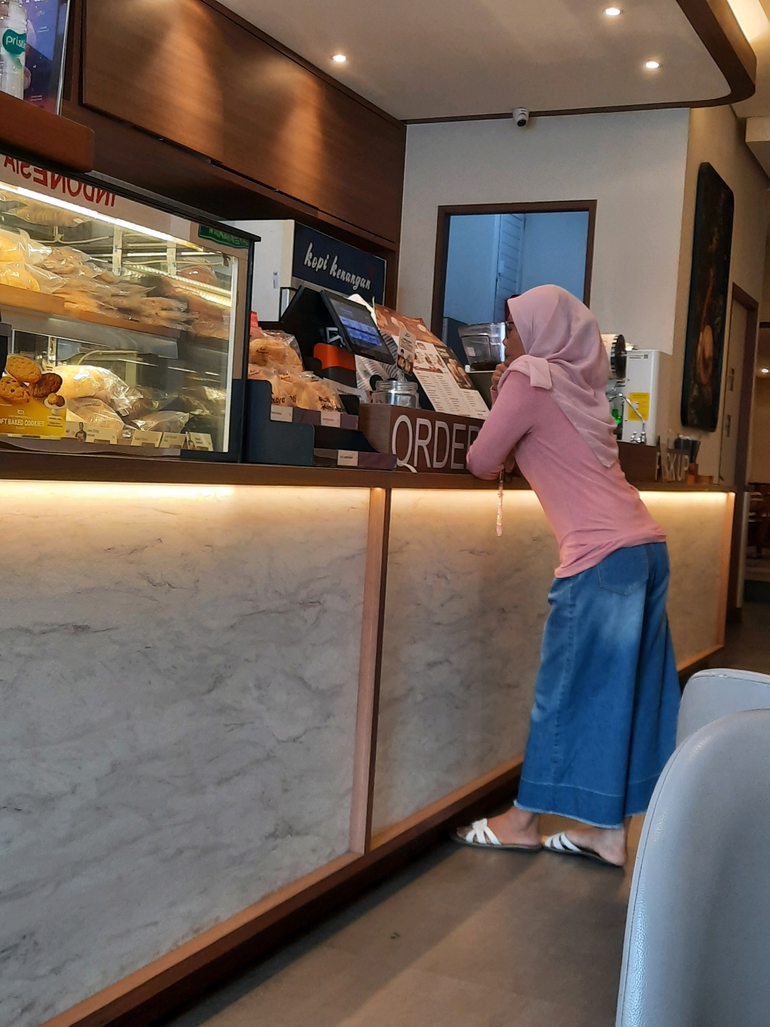 In East Jakarta, Indonesia, a woman wearing a pink hijab and a pink long-sleeved shirt with wide-leg blue jeans stands at a counter in what appears to be a cafe or bakery. She is leaning on the counter, looking towards the right of the frame. The counter features a display case filled with pastries and baked goods, and a point-of-sale system with a screen. A sign on the counter reads "ORDER." The environment is indoors with warm lighting, and a marble-patterned facade on the lower part of the counter. To the right, a camera is visible on the wall, and an artwork hangs on the wall.