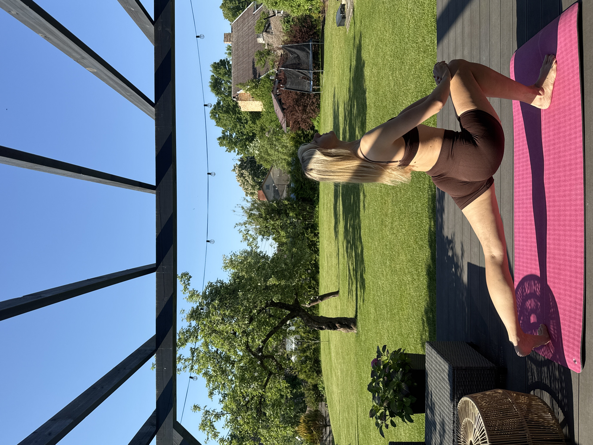 A portrait-oriented photograph captures a woman performing a yoga or stretching pose in an sunlit outdoor environment. The woman, with long blonde hair, is situated on the right side of the frame, facing left. She is dressed in a brown sports bra and matching shorts, kneeling on a bright pink yoga mat with one leg bent and foot raised, her hands clasped behind her head or neck in a deep stretch.
The yoga mat rests on a dark, grooved wooden deck, which transitions into a vibrant green grass lawn in the lower-left portion of the image. The lawn features long, distinct shadows, suggesting strong sunlight. In the midground, a large, leafy tree dominates the scene, and further back, residential houses and a trampoline are visible among more greenery.
The left side of the frame is characterized by prominent dark, angular wooden beams of a pergola structure, which extend across the top and down the side, framing a clear, brilliant blue sky. String lights are visible strung across the upper section of the yard, against the sky. The bright, cloudless sky and the sharply defined, elongated shadows on the grass and decking indicate a clear, sunny day, likely late morning or afternoon. Partially visible in the immediate foreground are outdoor wicker furniture and a dark planter with purple flowers. The scene depicts a serene and focused activity in a private garden setting in Carnikava, Latvia.