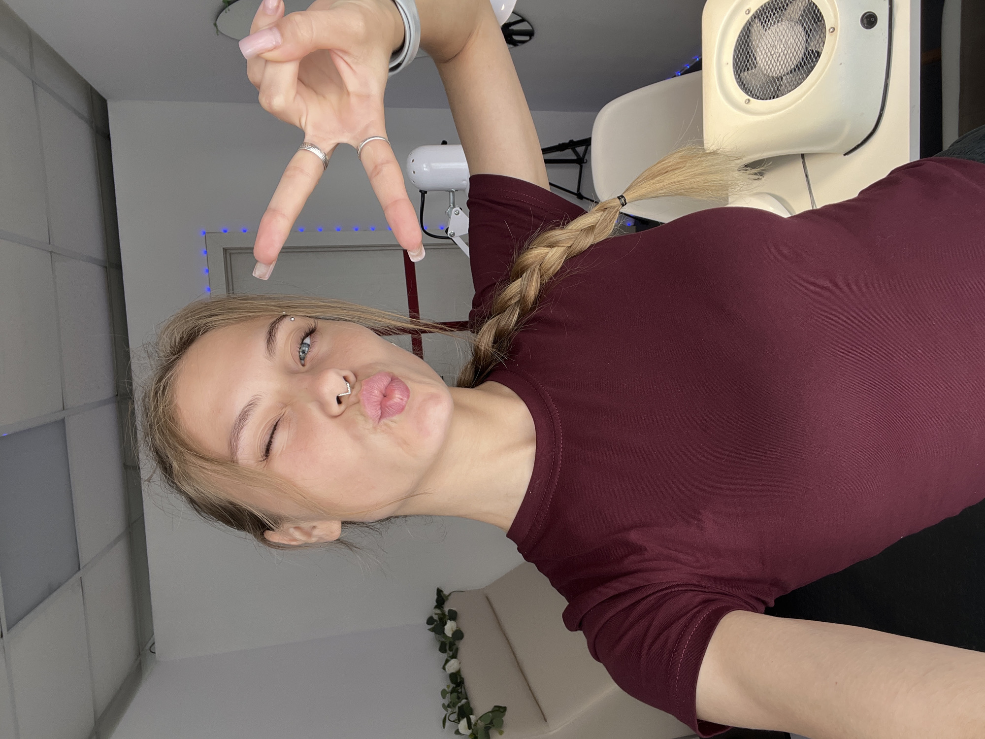 A young woman with blonde hair in a braid is posing for a selfie in a room that appears to be a nail salon. She is wearing a maroon t-shirt and has her right eye closed, her lips pursed, and her left hand raised in a peace sign. A septum piercing is visible on her nose. In the background, there is a white chair, a white lamp with a black base, and a white nail dust collector. The room is lit by overhead fluorescent lights and some blue fairy lights strung along the wall. The flooring is not visible. The location is not specified as being in the city of Фоки, Russia, and there is no information to support that claim.
