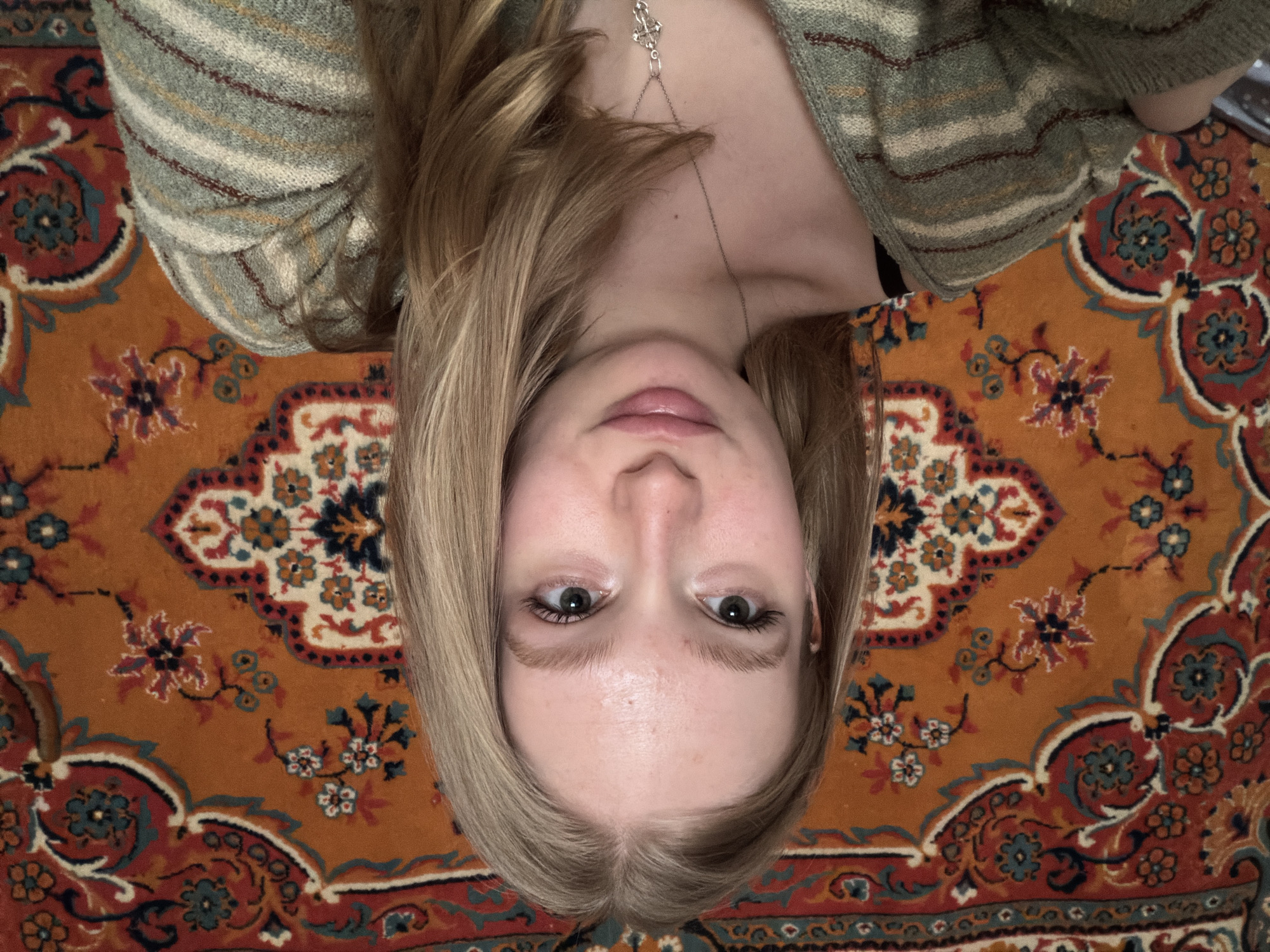 The image captures a fair-skinned individual, appearing female, positioned on their back and looking directly upward into the camera, resulting in an inverted perspective of their face. Light brown to blonde hair frames their head, and they have open, dark eyes with a neutral to slightly curious expression. Their lips are naturally pink. A delicate silver-colored necklace with a small, intricate charm, possibly a Celtic knot, is visible on their chest.
The person is wearing a soft, striped sweater featuring horizontal bands of light green, grey, cream, thin reddish-brown, and yellow. They are lying on a vibrant, intricately patterned rug that serves as the background. The rug has a dominant burnt orange or rust-colored base, adorned with elaborate floral and curvilinear designs in shades of deep red, various blues (including teal and navy), cream, and black.
The lighting is soft and even, suggesting an indoor setting with diffused light. There are no harsh shadows, and the overall mood is casual and relaxed. No visible text appears in the image. The location context is Засечное, Russia.