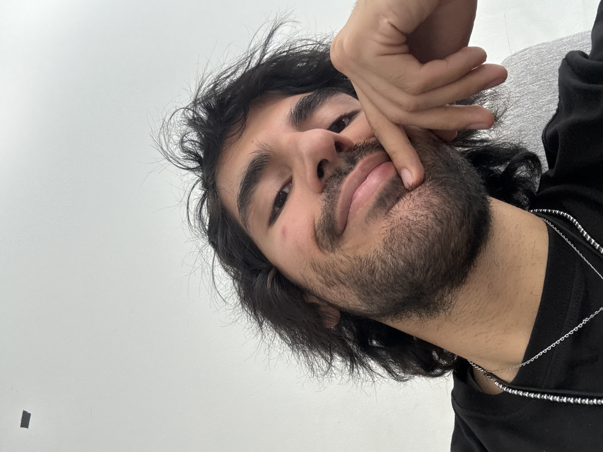 A close-up selfie of a man with dark hair and a beard. He is wearing a black shirt and a silver chain necklace. His right hand is near his face, with his index finger touching his cheek. The background is a plain, light-colored wall. The photograph appears to have been taken indoors under diffused lighting. The location is not discernible from the visual information provided. Yerevan, Armenia cannot be determined from this image.