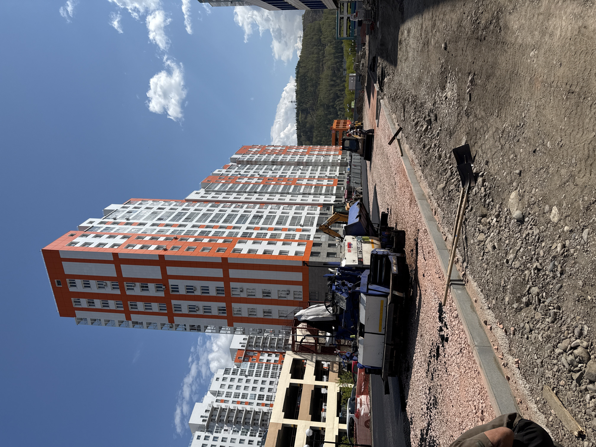 The image depicts a construction site adjacent to newly constructed apartment buildings in Krasnoyarsk, Russia. The buildings are high-rise, with orange and white facades, under a sky with scattered clouds. Construction machinery is visible, including a paver laying red gravel for a sidewalk. Shovels and other hand tools are present along the construction area. In the background, a wooded hill can be seen. The overall environment suggests an active construction zone on the outskirts of the city.