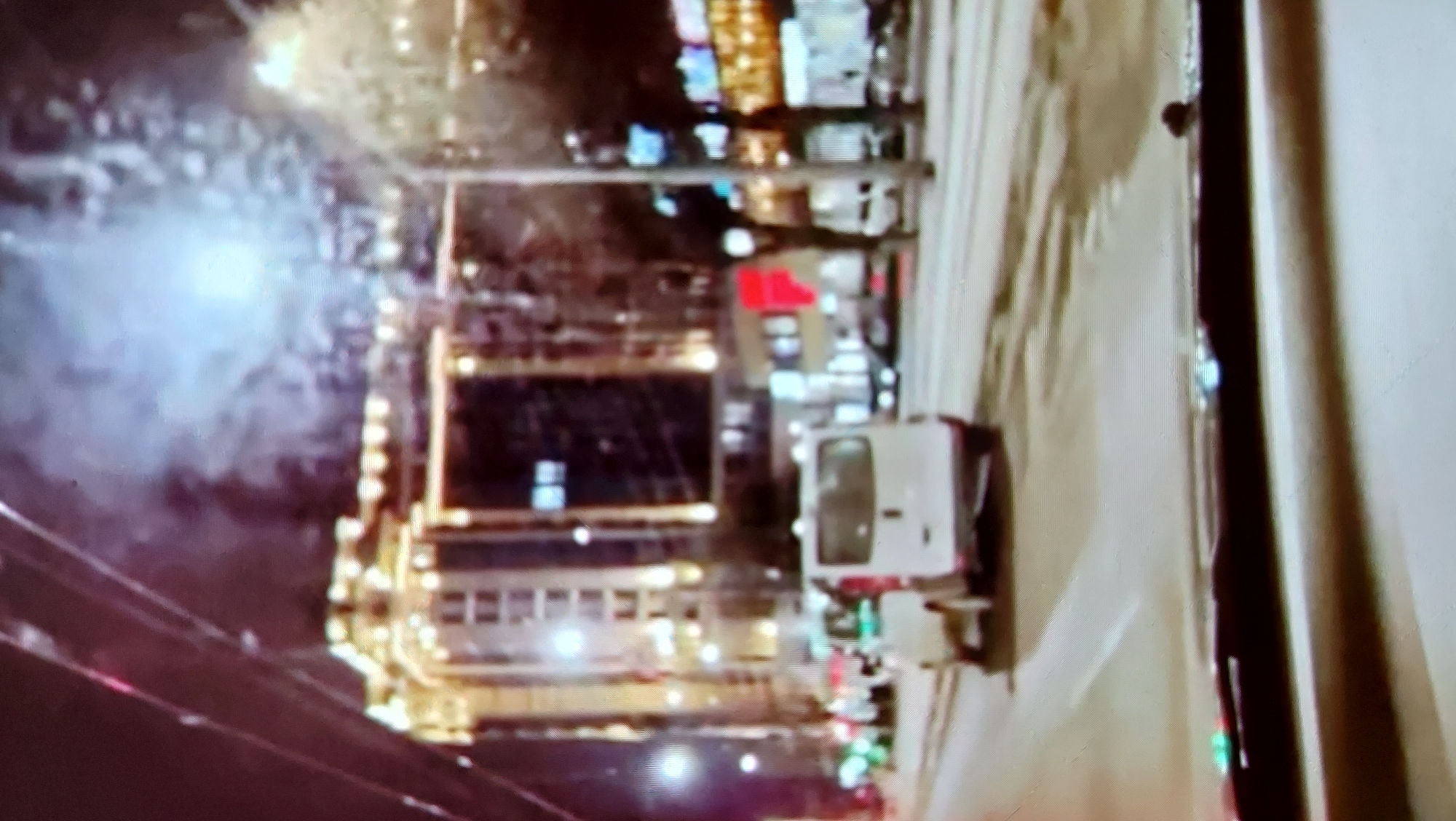 A nighttime city scene captured through a vehicle's windshield shows a white van driving on a road with city buildings and lights in the background. The van is white with a rear cargo door and appears to be moving forward. In the distance, a brightly lit building with multiple floors and illuminated windows dominates the skyline. Other vehicles, including a smaller white car, are also present on the road. The scene is characterized by a hazy, slightly blurred quality, suggesting motion or low light conditions. The overall impression is of a typical urban environment at night.