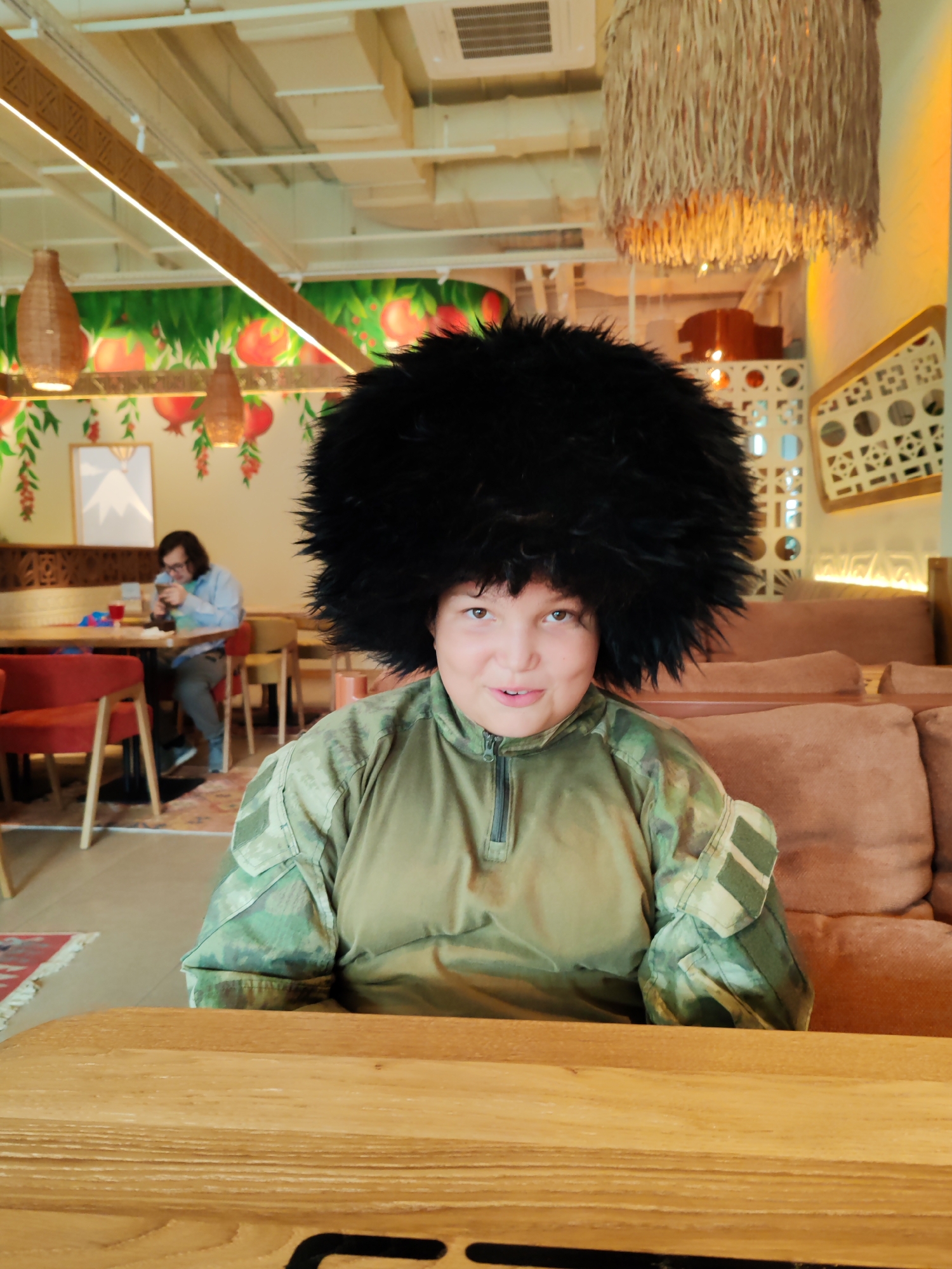 A person wearing a large, fuzzy black hat and a camouflage jacket sits at a wooden table in what appears to be a cafe or restaurant. The person is looking directly at the camera with a slight smile. Behind the person, a second individual is seated at another table, appearing to be looking at a phone. The environment features decorative elements such as hanging lamps, murals of pomegranates, and patterned wall decor. The lighting is warm and ambient. The overall setting suggests an indoor public space.