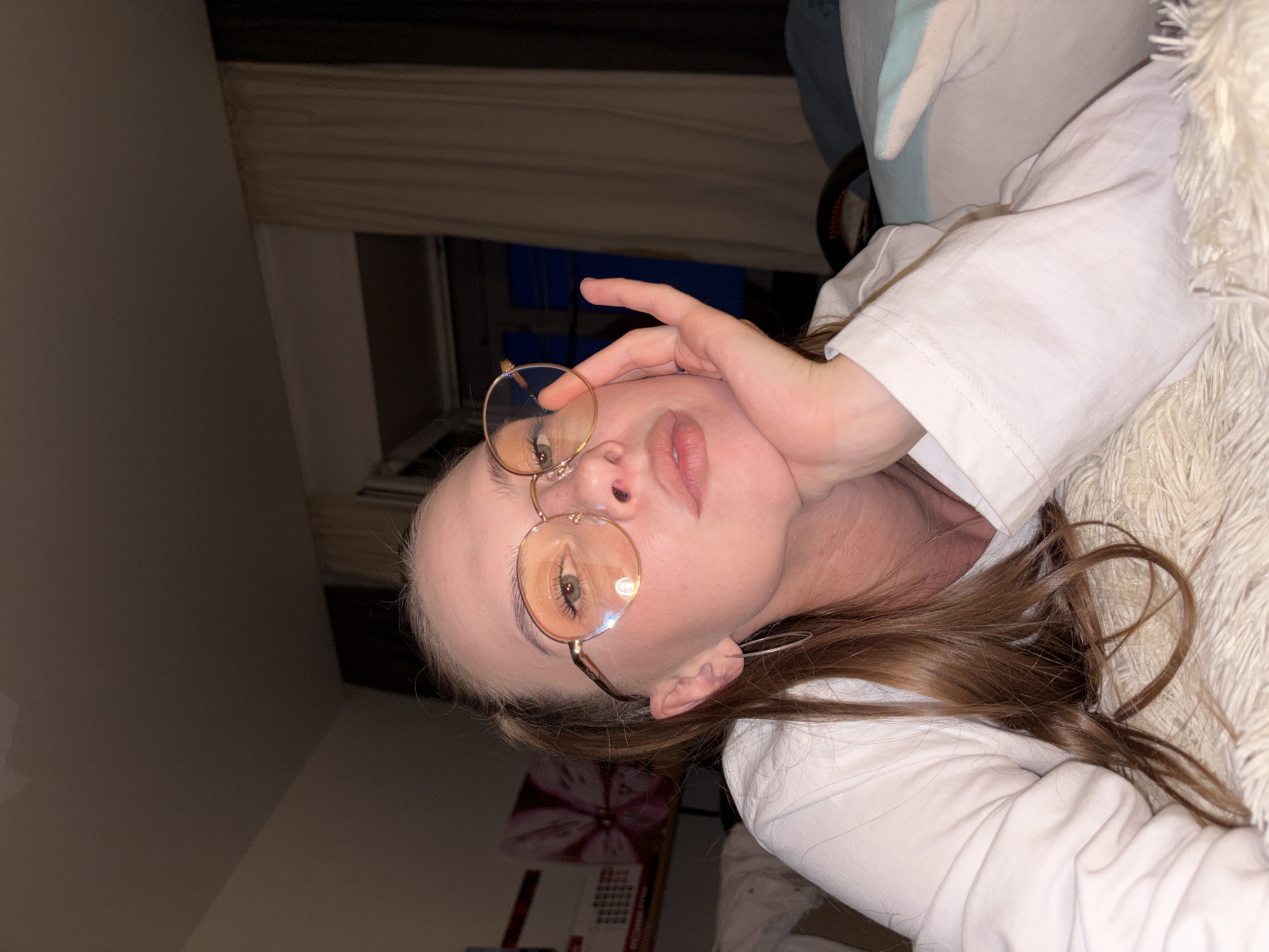 A young woman with light brown hair tied in a ponytail and wearing round, gold-rimmed glasses with orange lenses is lying on her side on a white, fluffy surface, possibly a bed or a large rug. She is looking towards the camera, resting her chin on her hand, with her lips slightly parted. She is wearing a white long-sleeved shirt. In the background, there are cream-colored curtains and a dark window, suggesting an indoor setting, likely a bedroom. The lighting is dim, with warm tones illuminating the woman's face and hair. There are no identifiable landmarks or objects that would indicate the city of Perm, Russia.