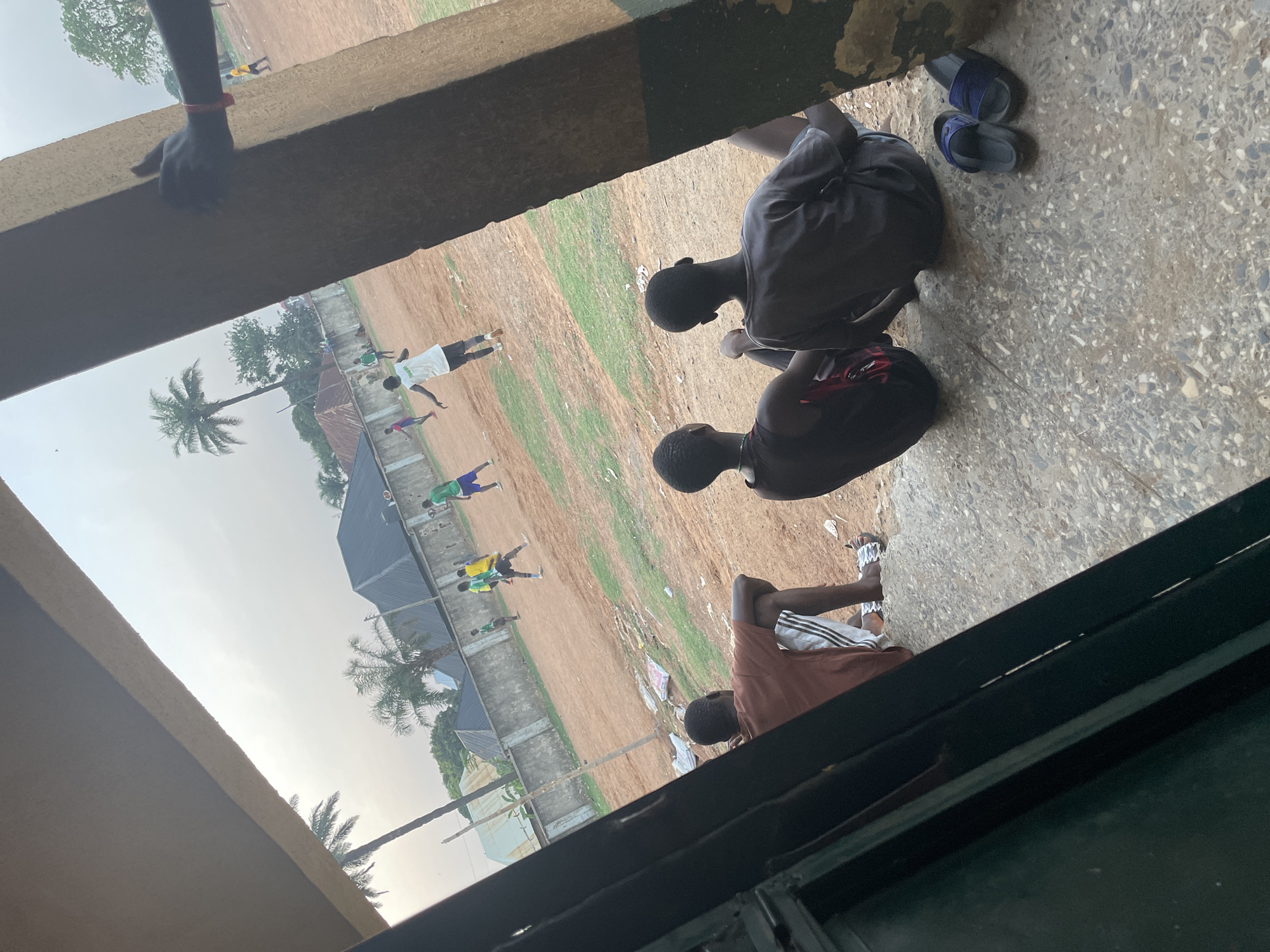 The image captures an outdoor scene in Akwanga, Nigeria, viewed from inside a building or shaded area. The perspective looks out over a dusty, open field bordered by simple buildings and palm trees.
In the immediate foreground, a dark-skinned hand, adorned with a red bracelet, rests on a concrete ledge. Below this ledge, on a patterned concrete floor, several young males are seated. To the right, two boys are seen from behind, wearing dark t-shirts and shorts. Beside them, a pair of blue sandals are placed on the floor. Further to the right, another young male, also seated, wears a brown t-shirt and white shorts with black stripes, resembling Adidas sportswear. A dark backpack is visible near him.
Beyond the foreground structure, a wide, reddish-brown dirt field dominates the mid-ground. Patches of green grass are interspersed throughout the field. On this field, several individuals, mostly young males, are active. One person in a white t-shirt and dark shorts is visible, while others further back wear green, yellow, and blue shirts, appearing to be playing or exercising. The field is lined with low fences and various buildings with dark corrugated iron roofs. Numerous palm trees and other vegetation are scattered across the landscape.
The sky is bright and hazy, suggesting it is daytime, possibly late afternoon or early morning. The overall atmosphere appears casual and relaxed, with some individuals observing the outdoor activities from a shaded vantage point. No distinct text is clearly legible on any clothing or objects.