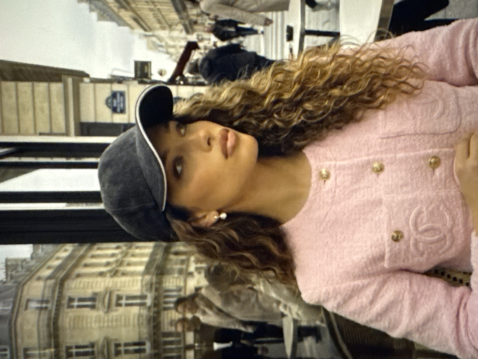 The image captures a close-up of a woman with curly brown hair wearing a pink tweed jacket with gold buttons and a black baseball cap. She is seated at a table, and in the background, several people are walking on a street with classic European-style buildings. The scene appears to be an outdoor cafe. The location is identified as the city of Kaunas, Lithuania.