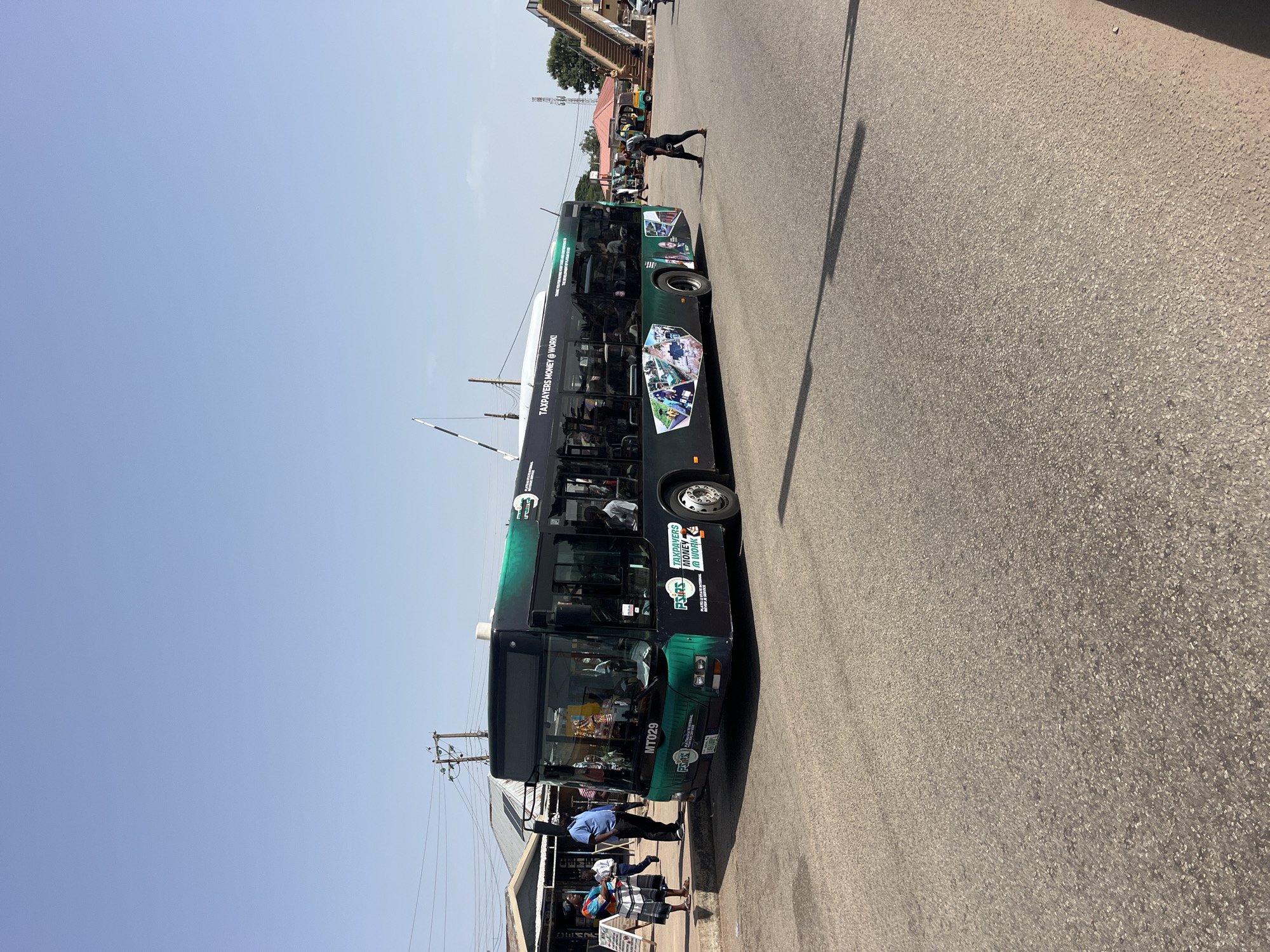 A wide shot captures a bus in motion, on a paved road in a city. The bus is predominantly black with green accents and graphics. The text on the bus reads "TAXPAYERS MONEY @ WORK" and "PLATEAU STATE INTERNAL REVENUE". Several passengers are visible through the tinted windows of the bus. A group of people are standing on a sidewalk to the left of the bus, including a woman in a patterned skirt and top and a man in a blue shirt. Another person, a woman in a black outfit, is walking across the road in front of the bus. In the background, there are buildings, power lines, and a pedestrian overpass with stairs. The sky is clear and blue, indicating daytime.