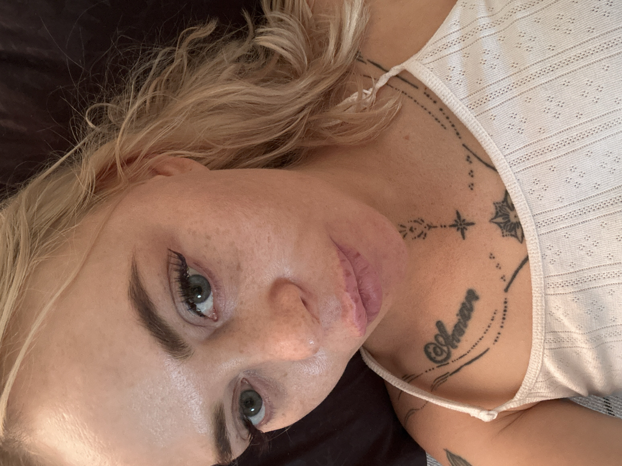 A close-up, high-angle selfie shows a young Caucasian woman with blonde hair and blue-green eyes. She is wearing a white tank top with a delicate, textured pattern. A black tattoo in cursive script is visible on her chest, reading "Annnan." Additional black line tattoos are present on her collarbone and shoulder area. The woman has light skin with visible freckles and is wearing mascara and eyeliner. The background is dark and appears to be a soft surface, possibly fabric.
