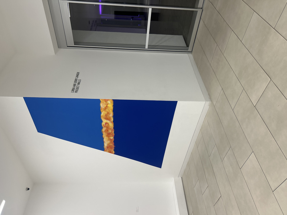 A brightly lit interior space displays a large, angular artwork. The piece features a vibrant blue background bisected by a vertical line of golden, textured material. "Linda and Jerry Janger Project Walls" is printed in small letters above the artwork. A doorway is visible in the upper portion of the frame, and a tiled floor extends along the right side. The photograph is taken at an angle, creating a dynamic perspective.