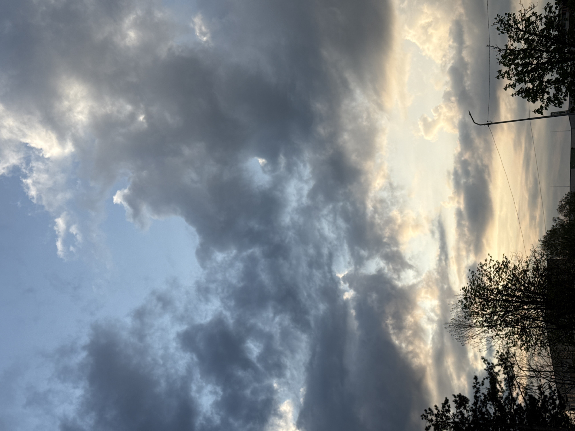 The image shows a sky with contrasting cloud formations. The left side of the frame features dark, heavy clouds, while the right side displays clouds that are brighter and lighter in color, possibly indicating the presence of sunlight. Trees are visible in silhouette on the right side of the image. A street lamp is also present on the right side of the image. This scene is likely located in an urban environment, specifically in the city of Pleven, Bulgaria.