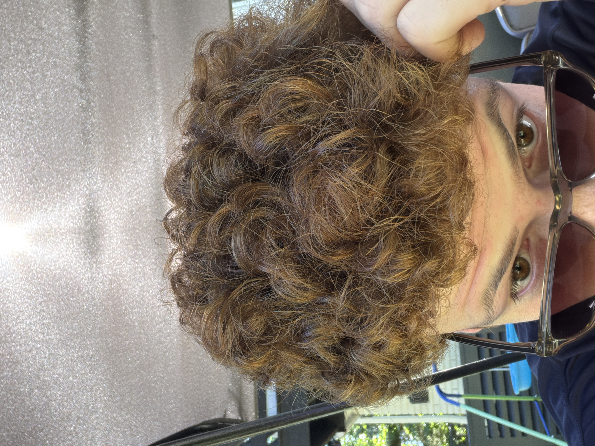 A close-up, eye-level shot captures the top of a person's head and their face, partially obscured by sunglasses. The individual has auburn, curly hair, and is wearing a dark shirt. They are outdoors, with a textured, light-colored canopy overhead and glimpses of greenery and outdoor equipment in the background. The lighting suggests it is daytime. The image appears to have been taken in Pepper Pike, United States of America.
