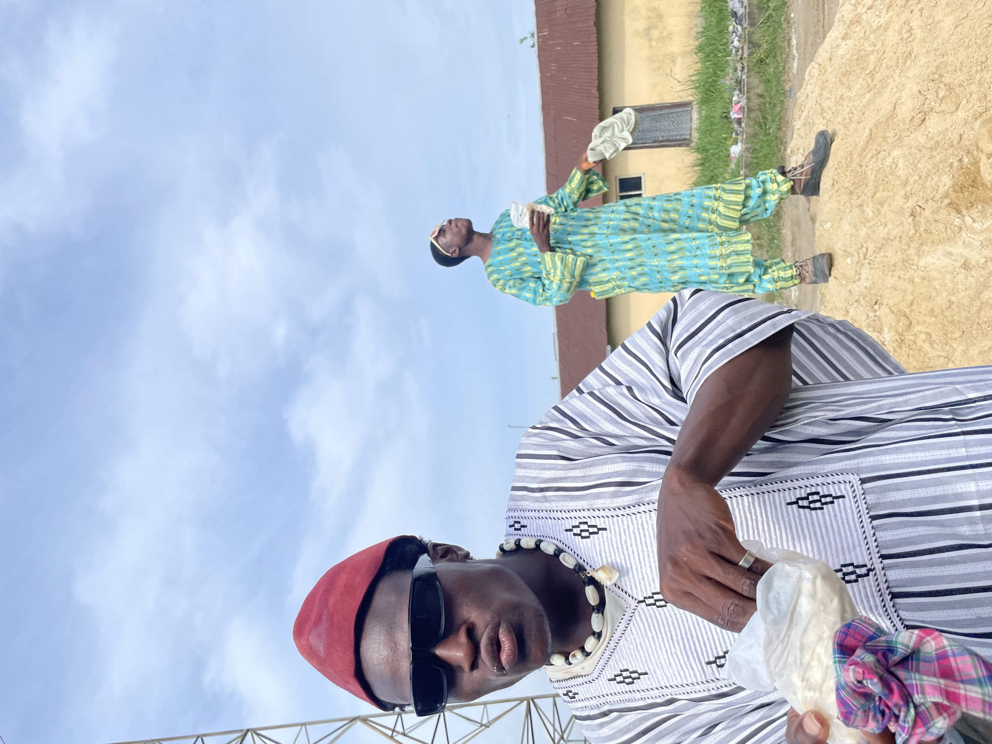 Two men are outdoors under a sky of pale blue streaked with white clouds. One man is in the foreground. He wears a red cap, sunglasses, a striped shirt, and a necklace of white beads. He is holding a piece of white cloth in one hand. The other man is further back, wearing a long, patterned, turquoise and yellow robe. He is looking upward and holds a piece of white cloth in his hand. There is a low, sloping pile of dirt or sand behind him. Behind that is a light yellow building and green vegetation. The lower left corner of the image shows the top of some metal construction.