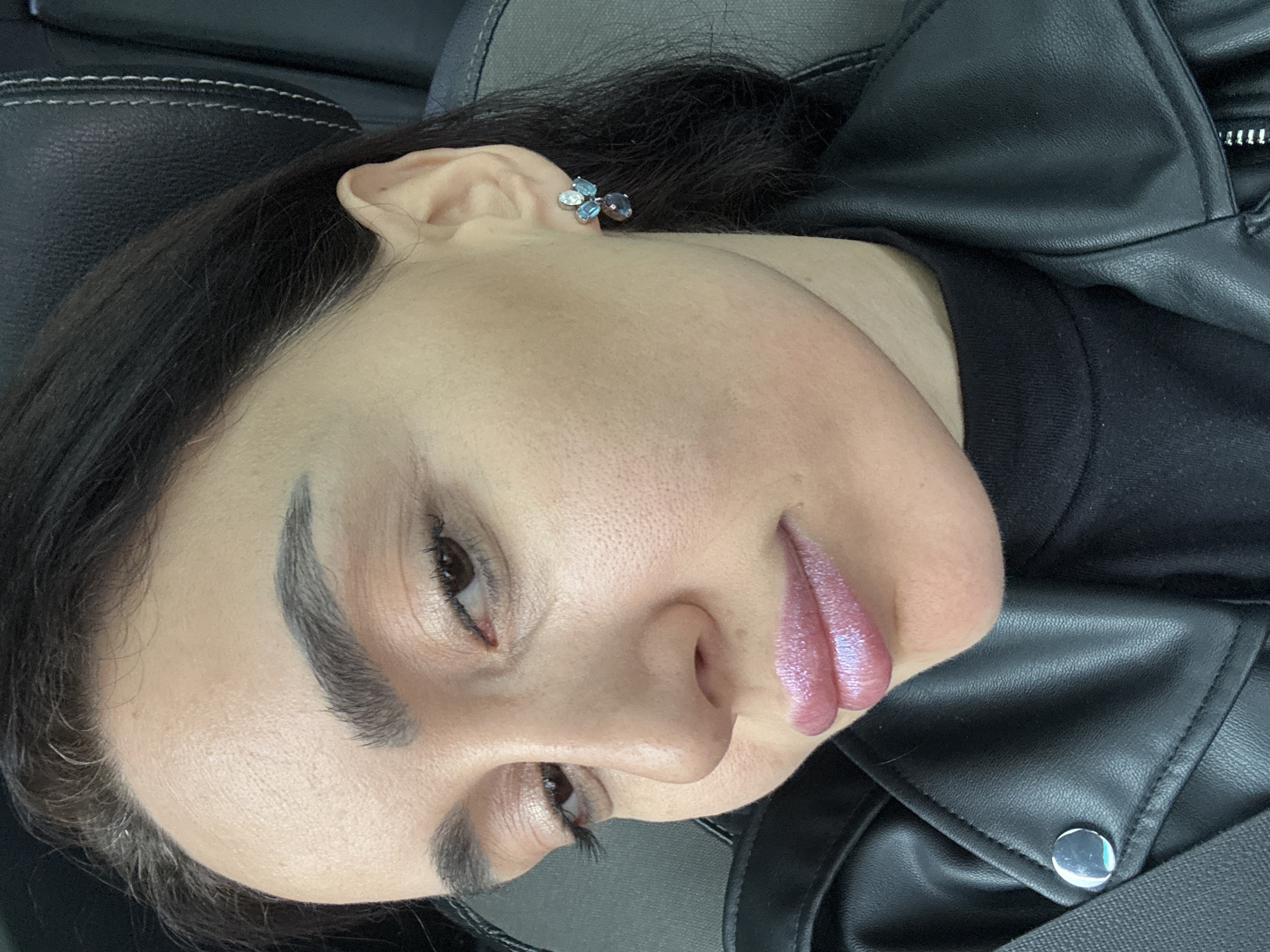 A close-up portrait shows a female subject from the upper chest, seated within the passenger compartment of a vehicle. The subject has dark hair, light skin, brown eyes with subtle eye makeup, and full, dark eyebrows. She wears shimmering pink lip gloss and a decorative earring on her visible right ear, consisting of small light-colored stones and a dangling darker teardrop-shaped stone. She is dressed in a black mock-neck garment under a black leather-look jacket with a prominent silver snap button on the collar. A black seatbelt strap crosses diagonally over her left shoulder/chest area. The background features a dark, textured vehicle seat headrest with light-colored stitching. The image contains no visual information to determine a specific location in Tbilisi, Georgia.