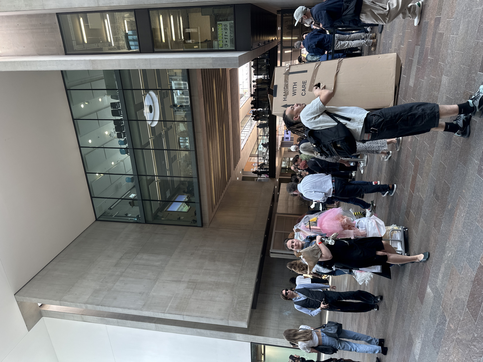 The image depicts a group of people in what appears to be the lobby or entrance hall of a large building. There are tall concrete pillars and walls. A large window reveals an office-like interior with chairs and lighting fixtures. A sign reading "WARNING 3-11-95" is partially visible on an upper level.
A line of people is visible in the background. In the foreground, people are standing and walking, some pulling carts and carrying items. One man in the center is carrying a large cardboard box with "HANDLE WITH CARE" printed on it, and seems to be taking a picture with his phone.
The scene takes place inside or near a public building in an urban setting.
It appears to be located in the Barbican Estate in the City of London, United Kingdom.