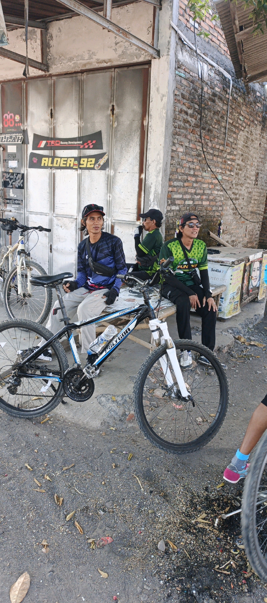 In Kebonan, Indonesia, three men are seated on a bench outside a storefront with a metal rolling door. The men appear to be resting. Two of the men are wearing cycling jerseys and hats, suggesting they may have been cycling. One man is wearing a blue and black long-sleeved shirt, grey pants, and a black fanny pack. Another man, seated to his right, is wearing a green cycling jersey with a black and white patterned collar and black pants, and he is holding a cigarette to his mouth. The third man, further to the right, is wearing a green cycling jersey with yellow accents, sunglasses, and black pants.
In front of the men are two bicycles. One is a black and blue mountain bike with the brand name "Polygon" visible on the frame and a water bottle attached to the frame. Another white bicycle is partially visible to the left of the frame. The storefront has several banners and signs displayed on the metal door, including one that reads "TRD Racing Development" and another with "ALDERA 92". The ground in front of the store is covered in dirt and debris, with some leaves scattered around. The wall of the building to the right is made of red brick. There are also some items displayed on a surface behind the men, which appear to be various products.