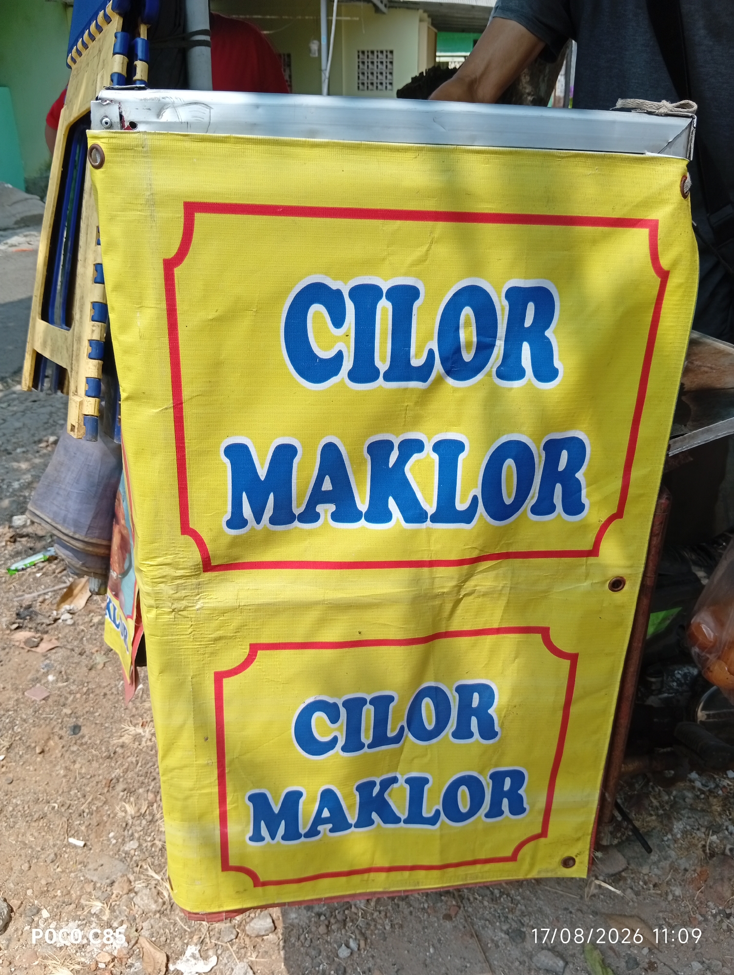 A yellow banner with red trim displays the text "CILOR MAKLOR" in large blue letters, repeated twice. The banner is attached to a cart with a metal frame. The cart appears to be a food stall, with various items visible in the background, including bags of goods. The ground is paved with dirt and rocks. The scene is outdoors, with sunlight casting shadows. The timestamp "17/08/2026 11:09" is visible in the lower right corner, suggesting this image was taken in South Tangerang, Indonesia. A person's arm is visible in the upper right portion of the image, and they appear to be operating the cart. The overall impression is that of a typical street food vendor in Indonesia.